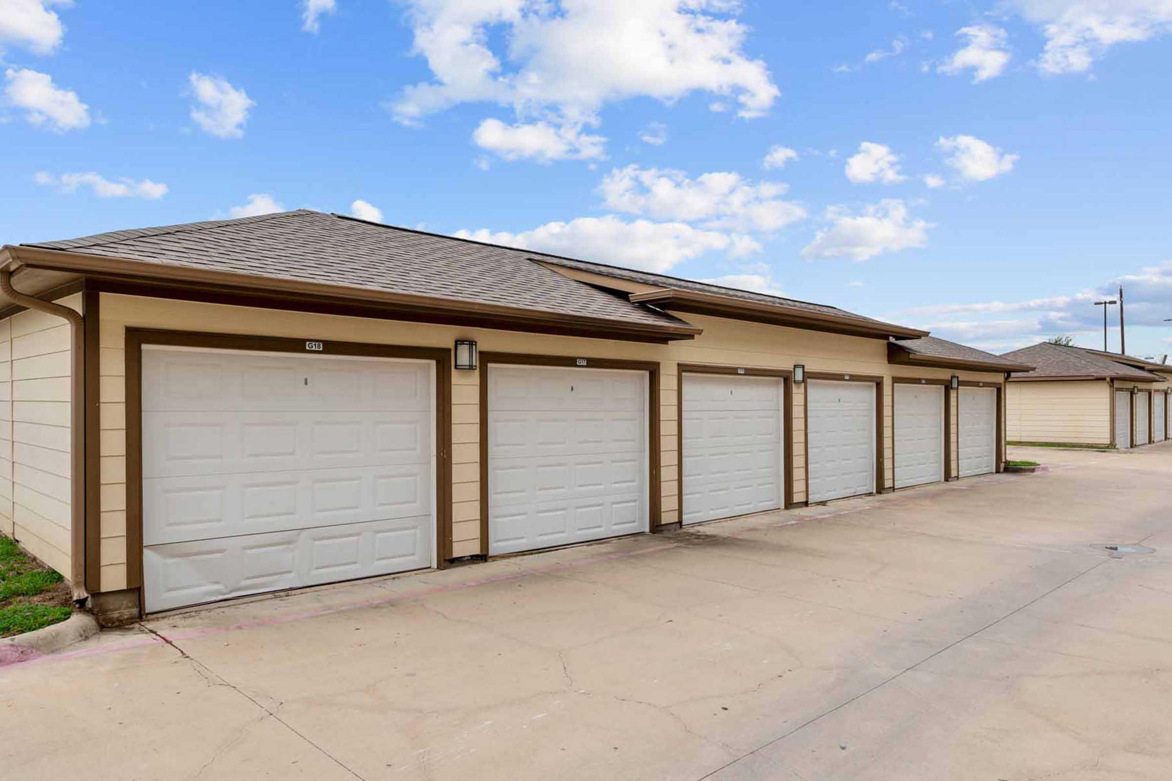 Row of six garage doors in a residential area, featuring white doors with panel designs and brown trim. The garages are lined up along a paved driveway, with green grass visible at the edges and a blue sky with scattered clouds above.
