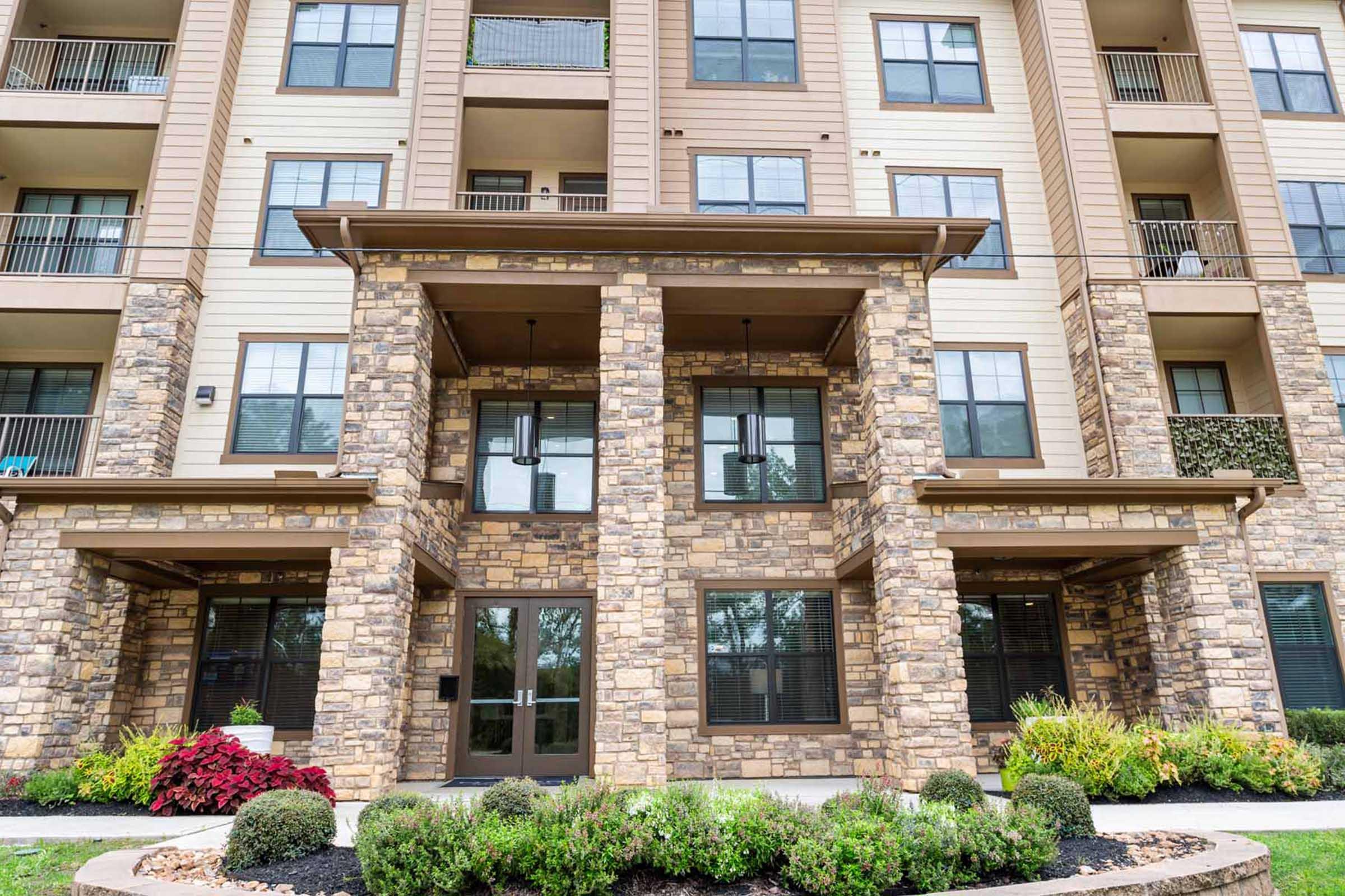 Exterior view of a multi-story apartment building featuring a stone facade, large windows, and a covered entrance. The landscape includes neatly trimmed shrubs and colorful flowers, creating an inviting atmosphere. Balconies are visible on the upper floors.