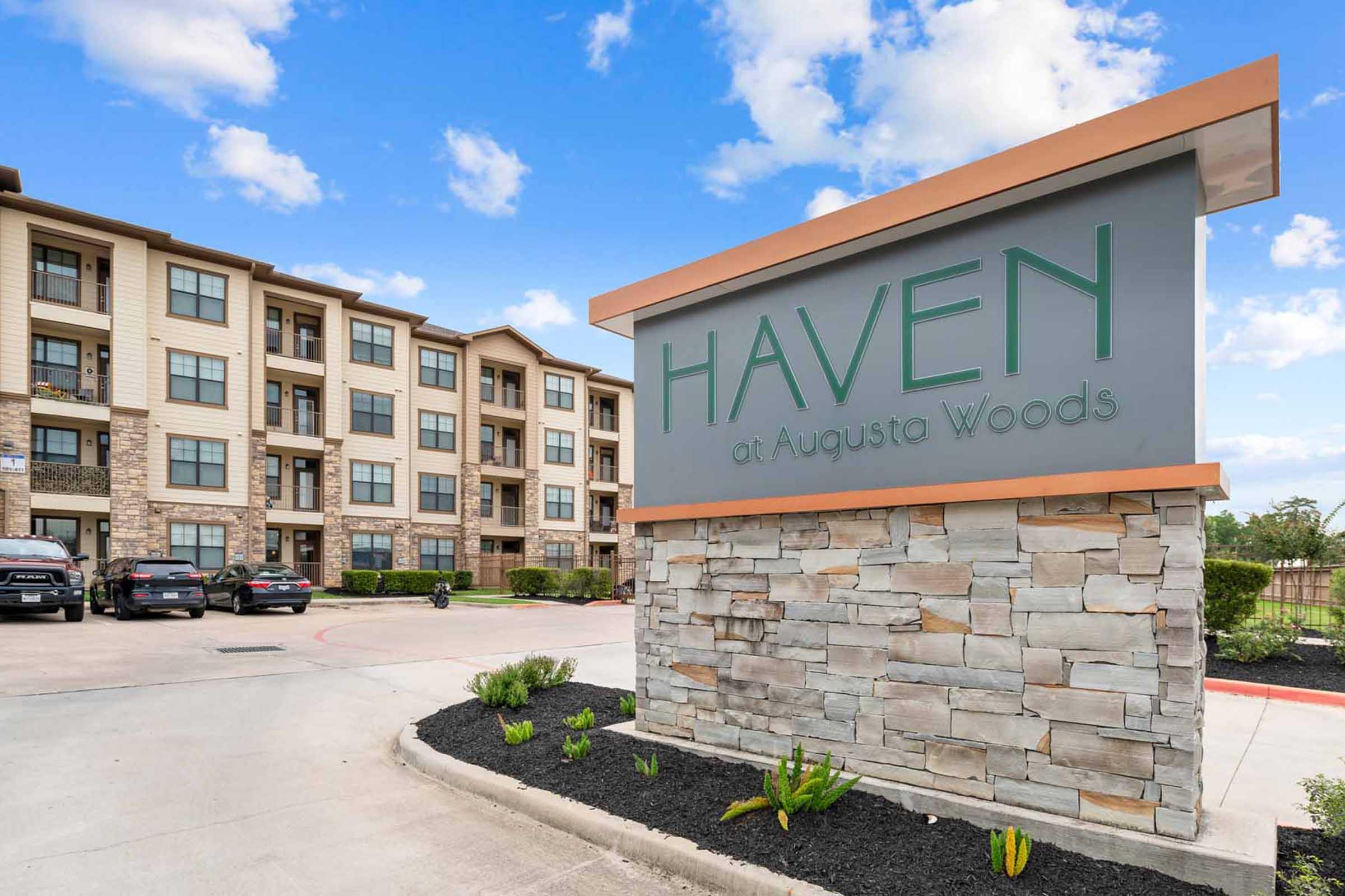Sign for "Haven at Augusta Woods" in front of a residential building. The sign features modern lettering and is mounted on a stone base. The background shows a well-maintained multi-story apartment complex with multiple windows, surrounded by landscaping and a clear blue sky.
