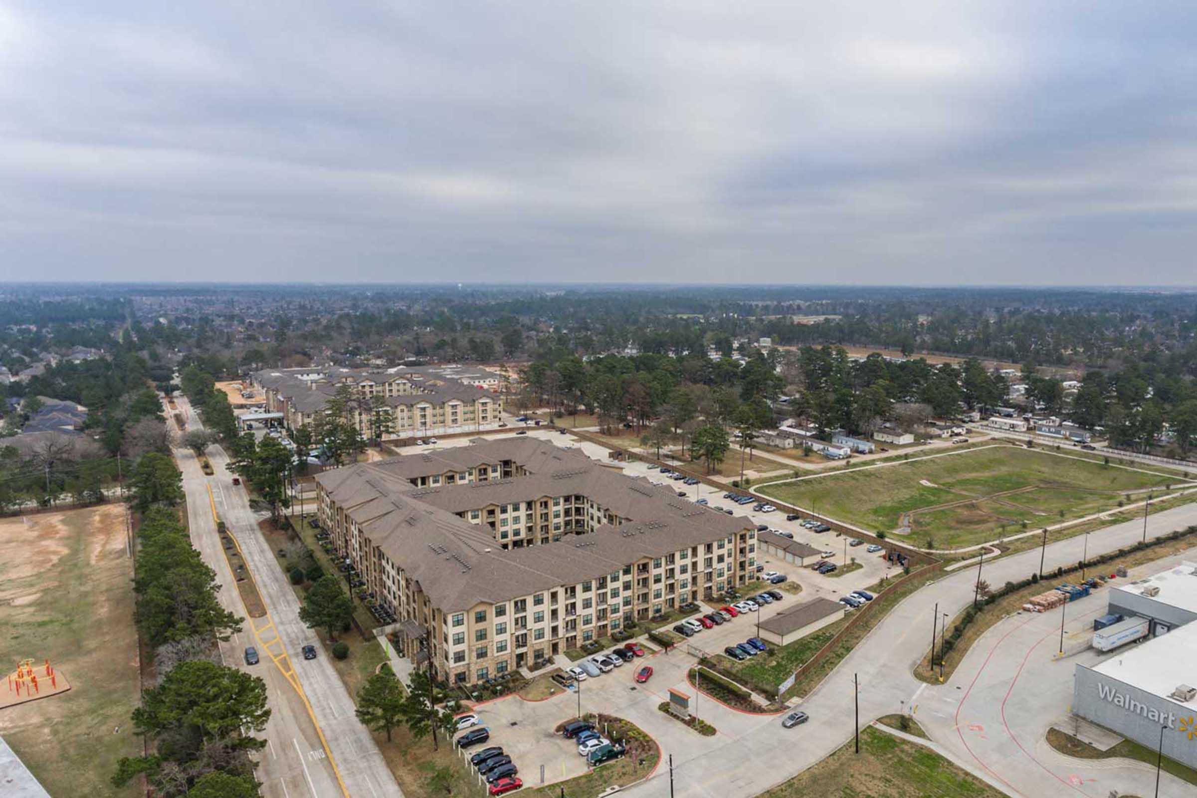 Aerial view of a suburban landscape showing a multi-story apartment complex surrounded by greenery and parking lots. Nearby structures include a Walmart and other commercial buildings, with a road running alongside. The sky is overcast, creating a muted atmosphere.