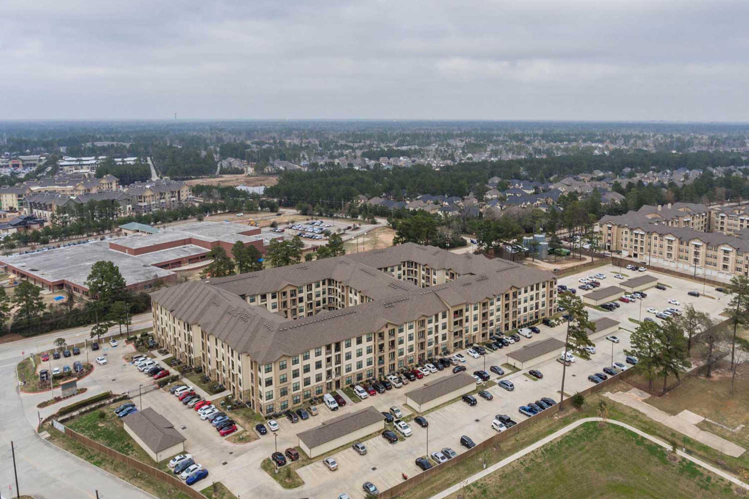 Aerial view of a multi-story apartment complex surrounded by greenery and a parking lot, with additional residential and commercial developments visible in the background. The scene includes a mix of buildings and open spaces under a cloudy sky.
