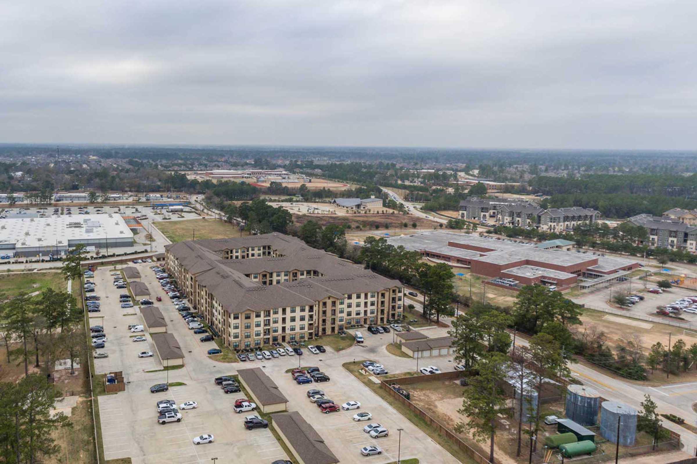 Aerial view of a large multi-story building surrounded by parking lots, with several cars visible. In the background, various construction sites and new developments are seen, including partially finished buildings and wooded areas. The sky is overcast, giving a muted atmosphere to the scene.