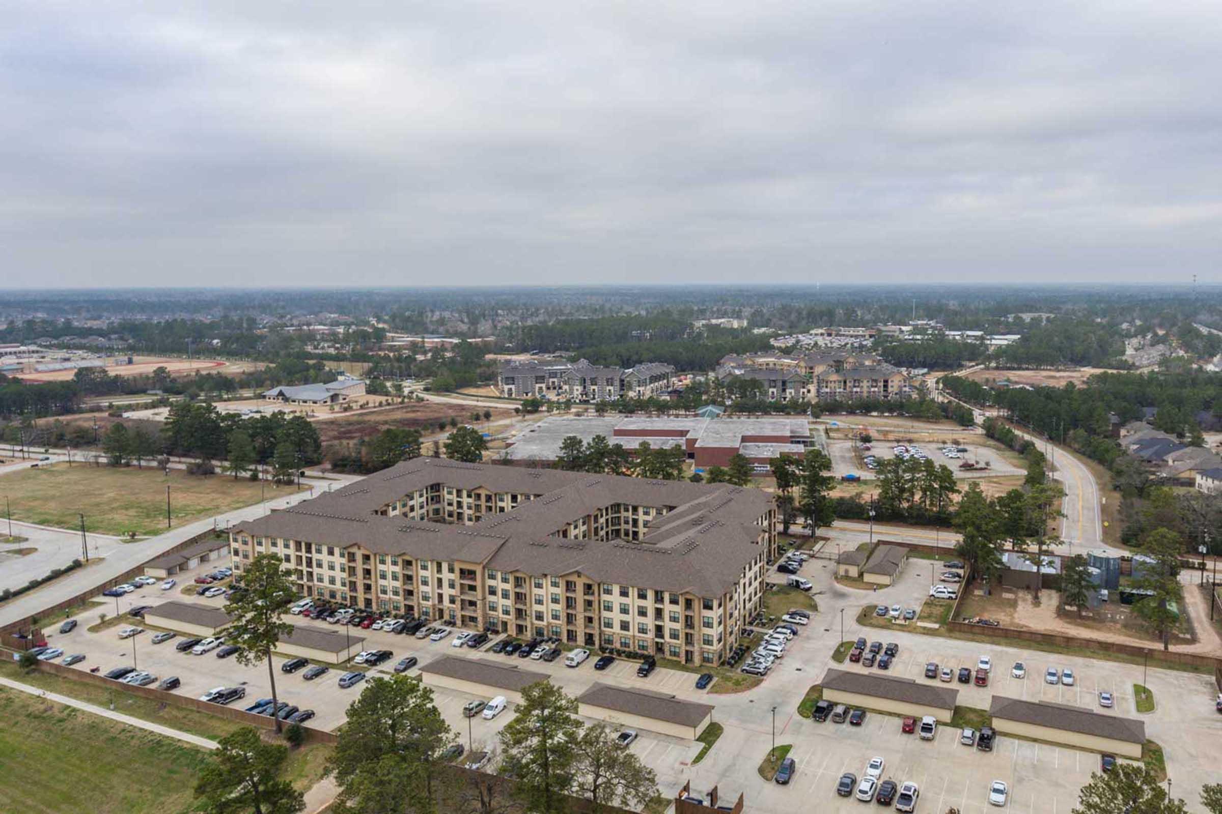 Aerial view of a multi-story apartment complex bordered by parking lots, with additional nearby buildings and green areas in the background. Overcast skies and a sprawling landscape featuring roads and other developments extend into the distance.