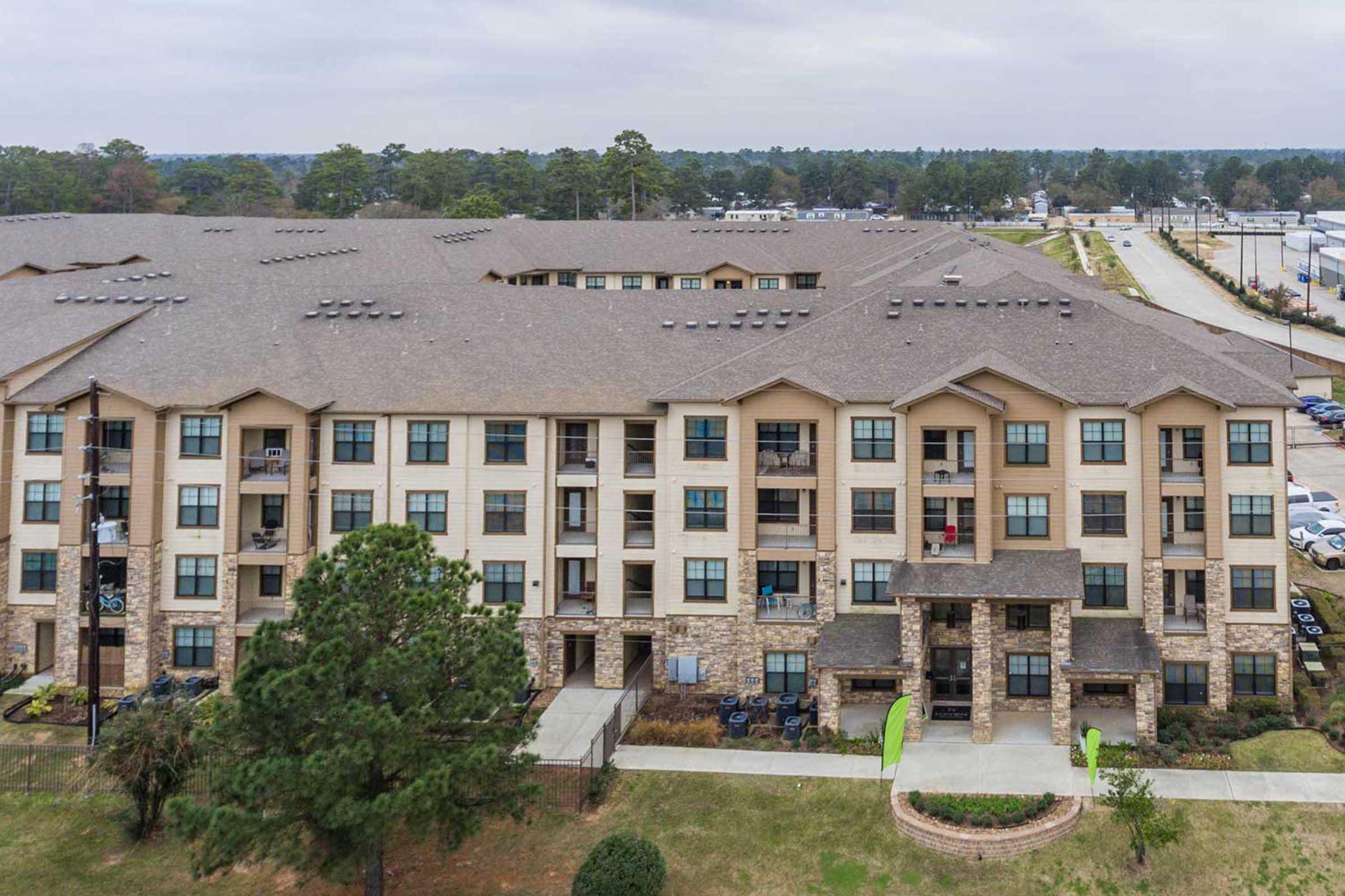 Aerial view of a multi-story apartment complex featuring a mix of stone and wooden exterior. The building has balconies and green accents, surrounded by grass and trees, with a road and additional buildings visible in the background. The sky is overcast.