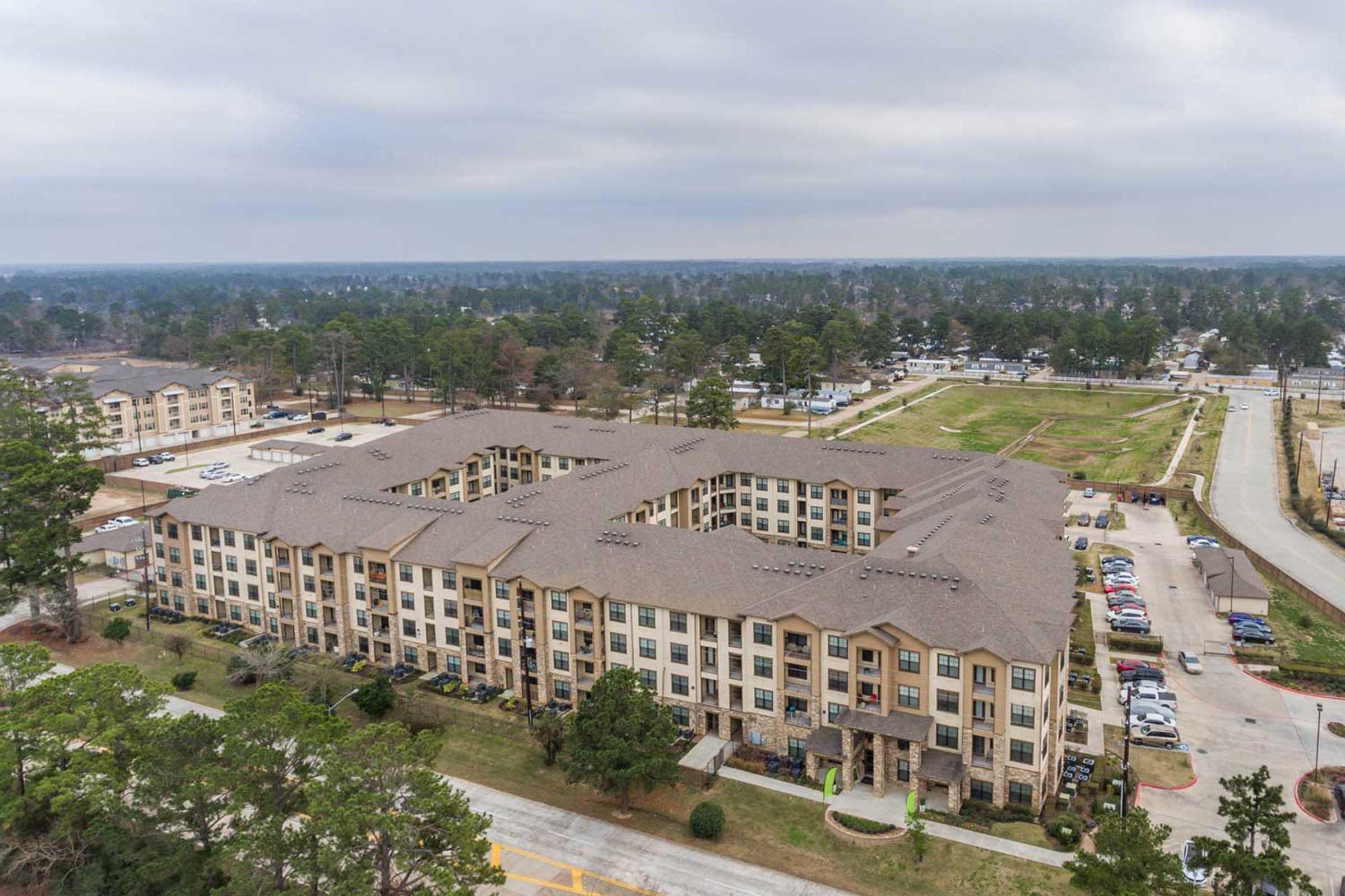 Aerial view of a multi-story apartment complex surrounded by greenery, with an urban area in the distance. The building features a mix of residential units and parking spaces, with pathways and landscaped areas visible. The sky is overcast, suggesting a cloudy day.