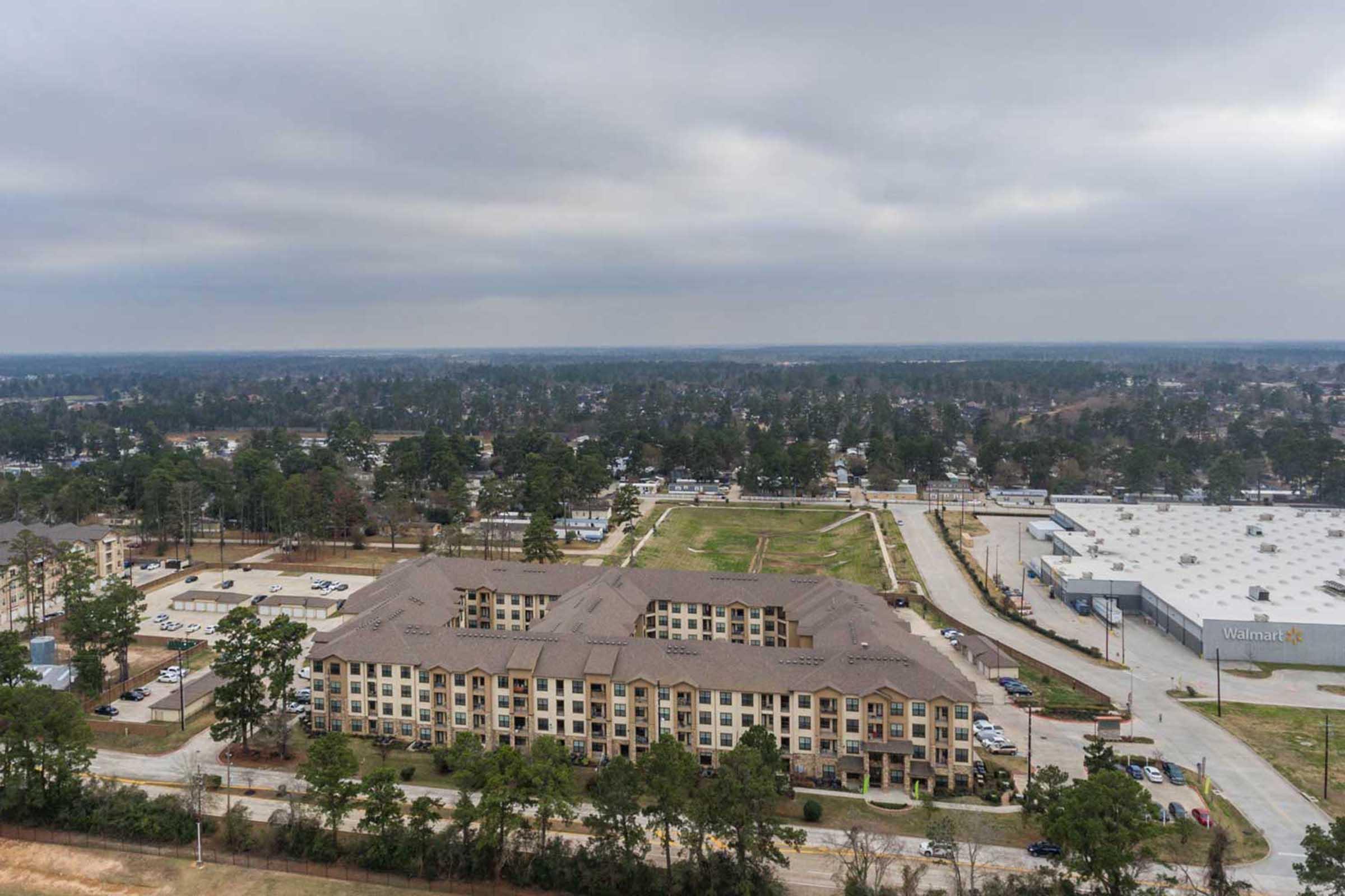 Aerial view of a residential building complex surrounded by trees, with a parking lot in the foreground. In the distance, a Walmart store is visible amid a suburban landscape under a cloudy sky. The scene captures a blend of greenery and urban development.