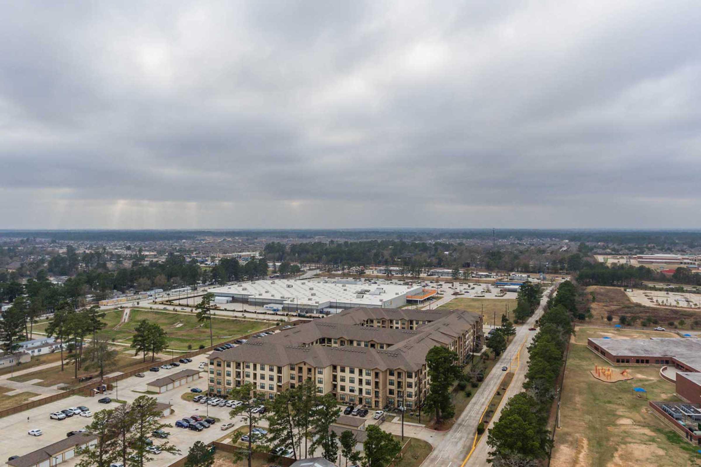 Aerial view of a multi-story residential building surrounded by parking lots and green areas, with a distant cityscape under a cloudy sky. The image captures a mix of urban and natural elements, showcasing a developing area with roads and trees.