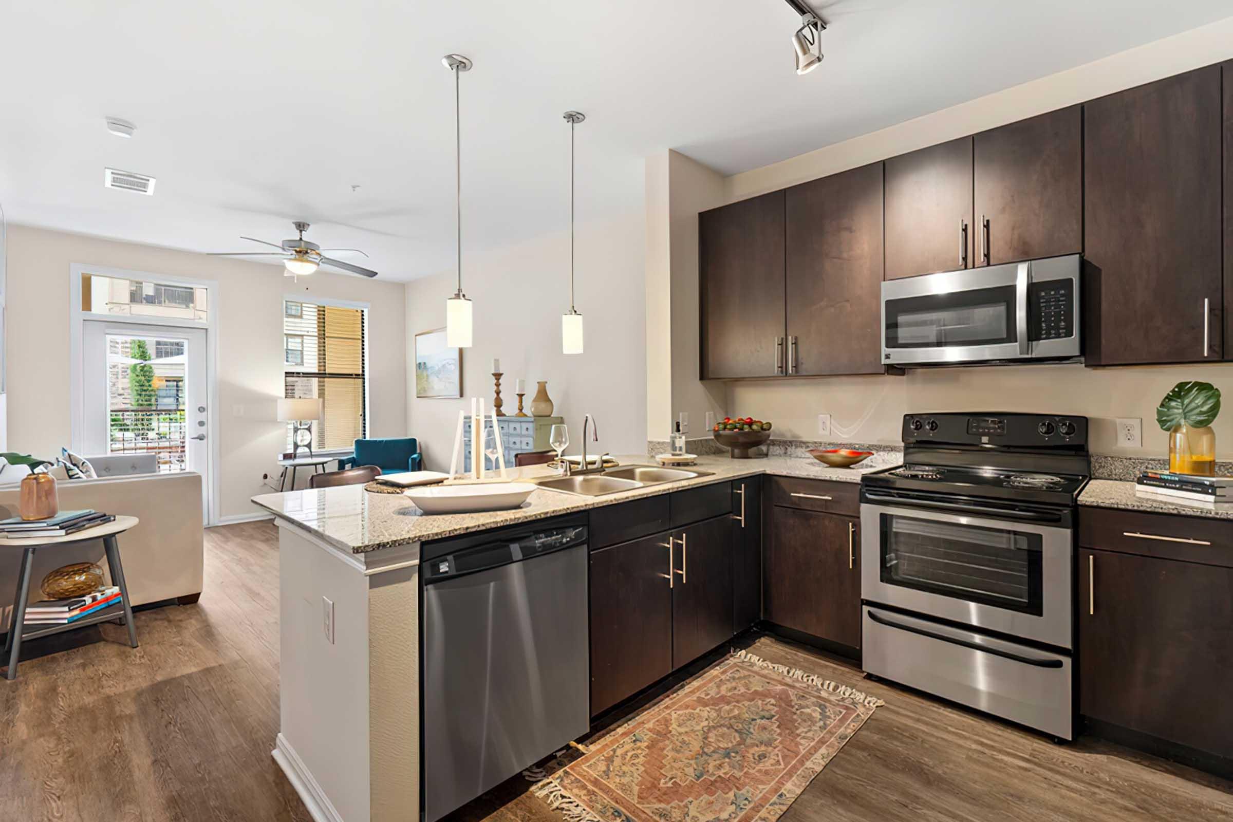 Modern kitchen featuring dark wooden cabinets, stainless steel appliances including a stove, microwave, and dishwasher. A granite countertop with a sink is visible, along with decorative items. In the background, a cozy living area can be seen through a window with natural lighting.