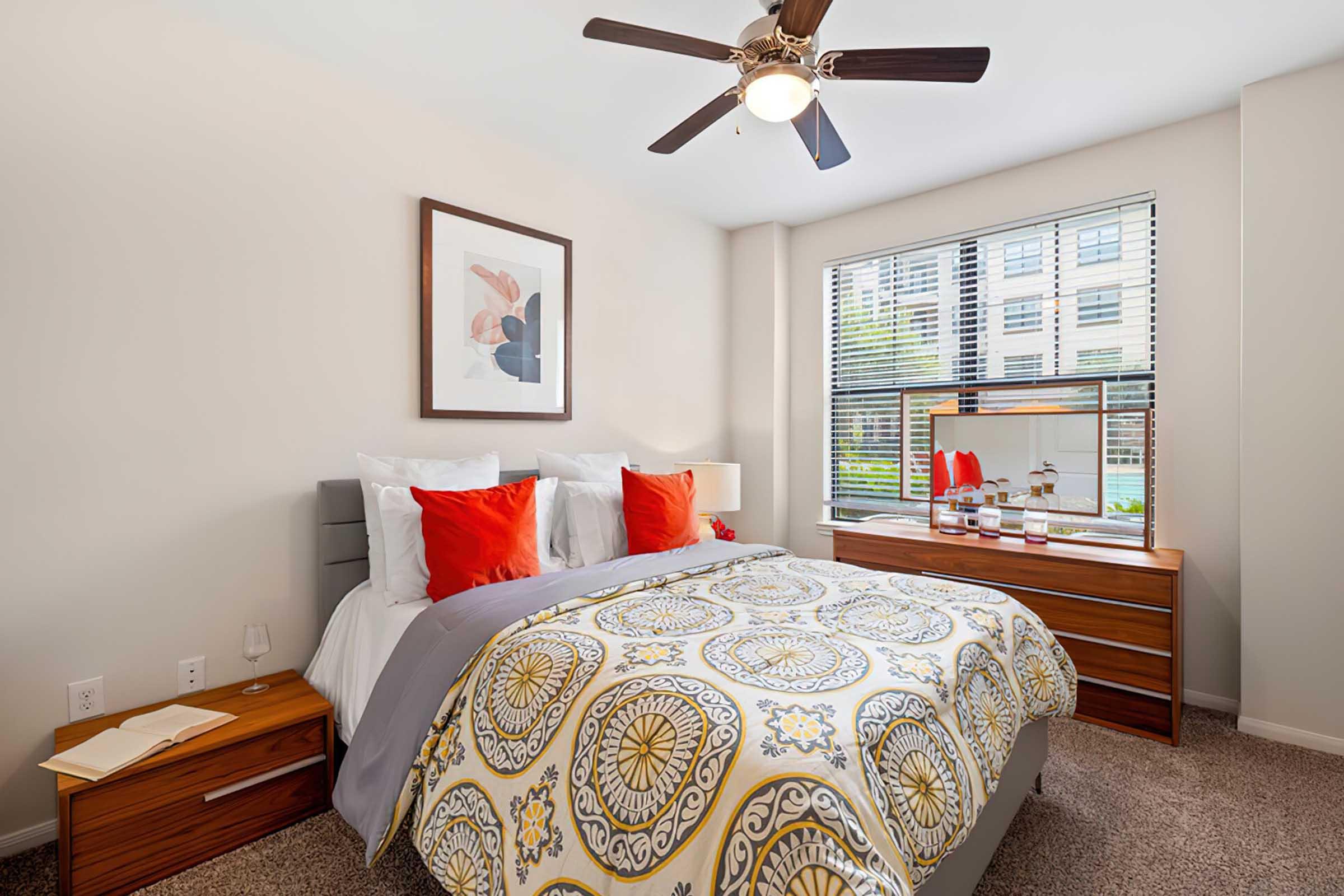 Bright and cozy bedroom featuring a queen-sized bed with a patterned yellow and gray comforter, red accent pillows, and matching nightstands. A modern ceiling fan provides ventilation, while large windows allow natural light to illuminate the space, complemented by a piece of abstract wall art and a decorative fish tank.