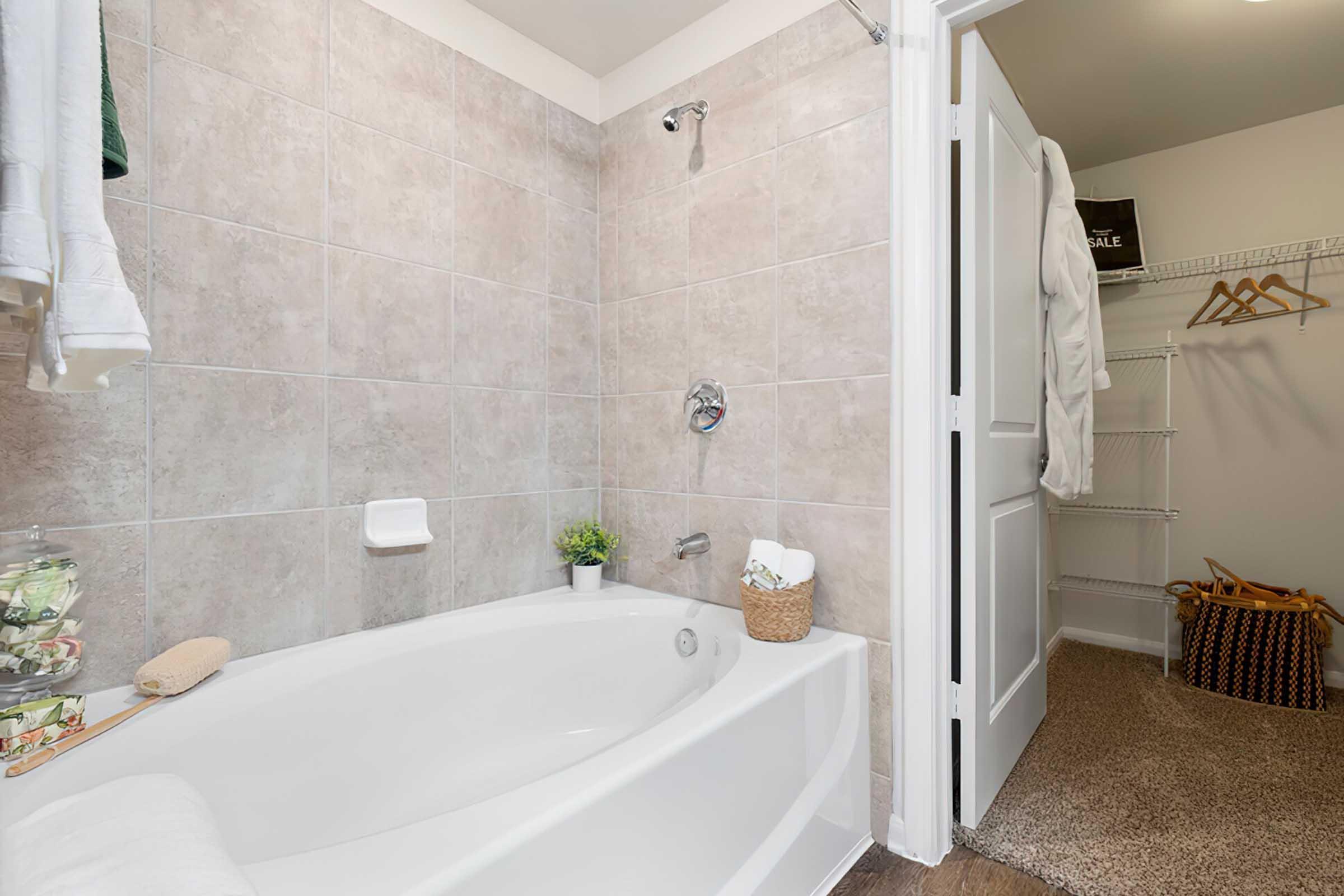 A clean bathroom featuring a white bathtub, tiled walls, a showerhead, and a towel rack. A small plant and toiletries are on the edge of the tub. A closet door is visible, with hangers and a basket in the closet. The floor is carpeted in brown tones.