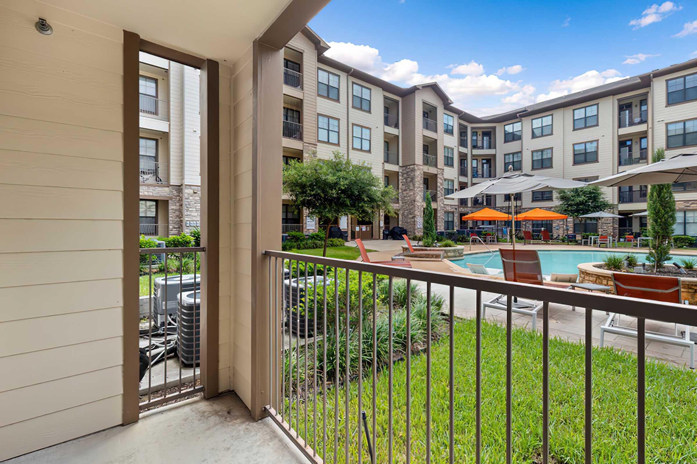 View from a balcony overlooking a well-maintained courtyard with a swimming pool. The area features lounge chairs and umbrellas, surrounded by greenery and residential buildings in the background. Bright blue sky adds to the inviting ambiance.