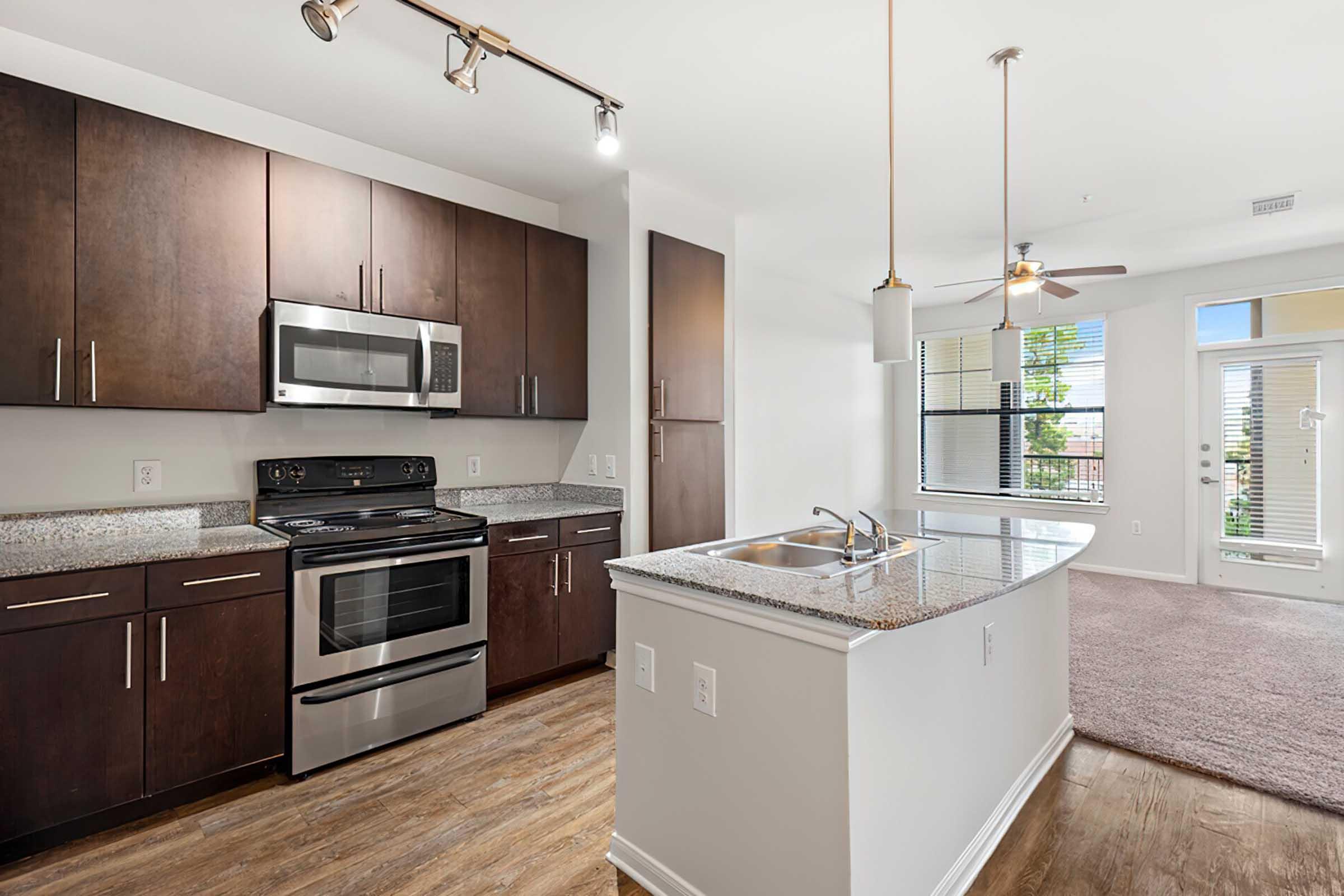 Modern kitchen with dark wooden cabinets, stainless steel appliances including a microwave and stove, and a granite countertop. The open layout features a living area with natural light pouring in through large windows. Light-colored walls and a ceiling fan complete the stylish design.