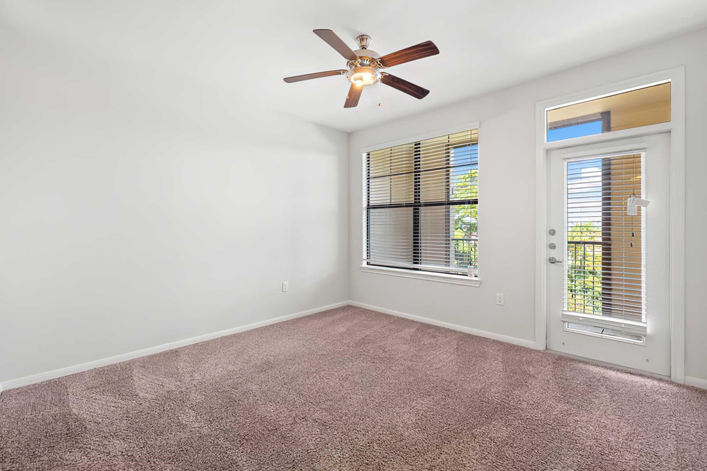 Spacious living room with light-colored walls, a ceiling fan, and large windows allowing natural light. Plush, pink carpet covers the floor. The room includes a door leading to a small balcony with a view.