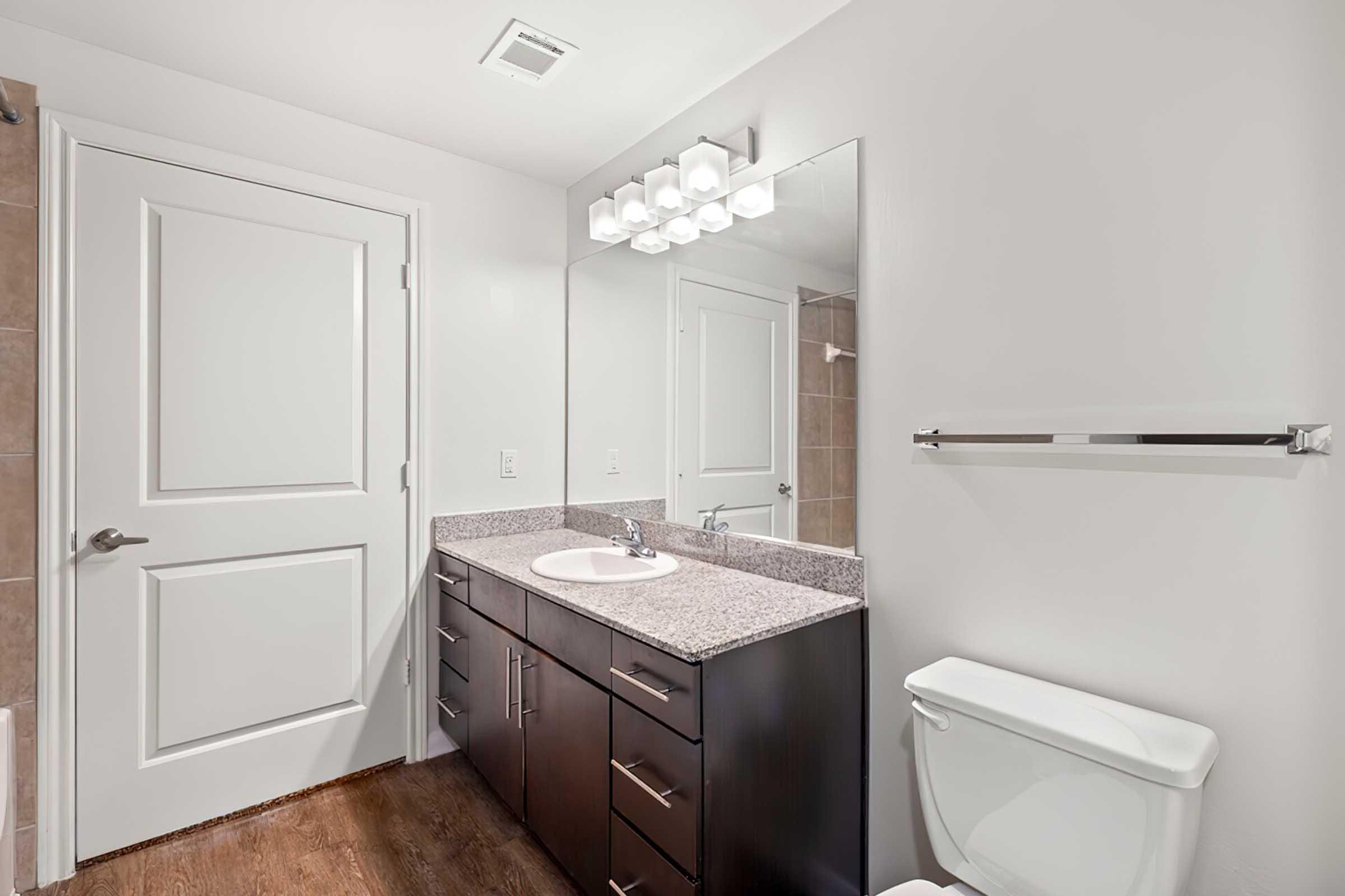 Modern bathroom featuring a white wall, a large mirror above a granite countertop with a sink, dark wood cabinets, a toilet, and a towel bar. The space is well-lit with ceiling lights, and there is a door visible leading outside the bathroom. The flooring is wooden.