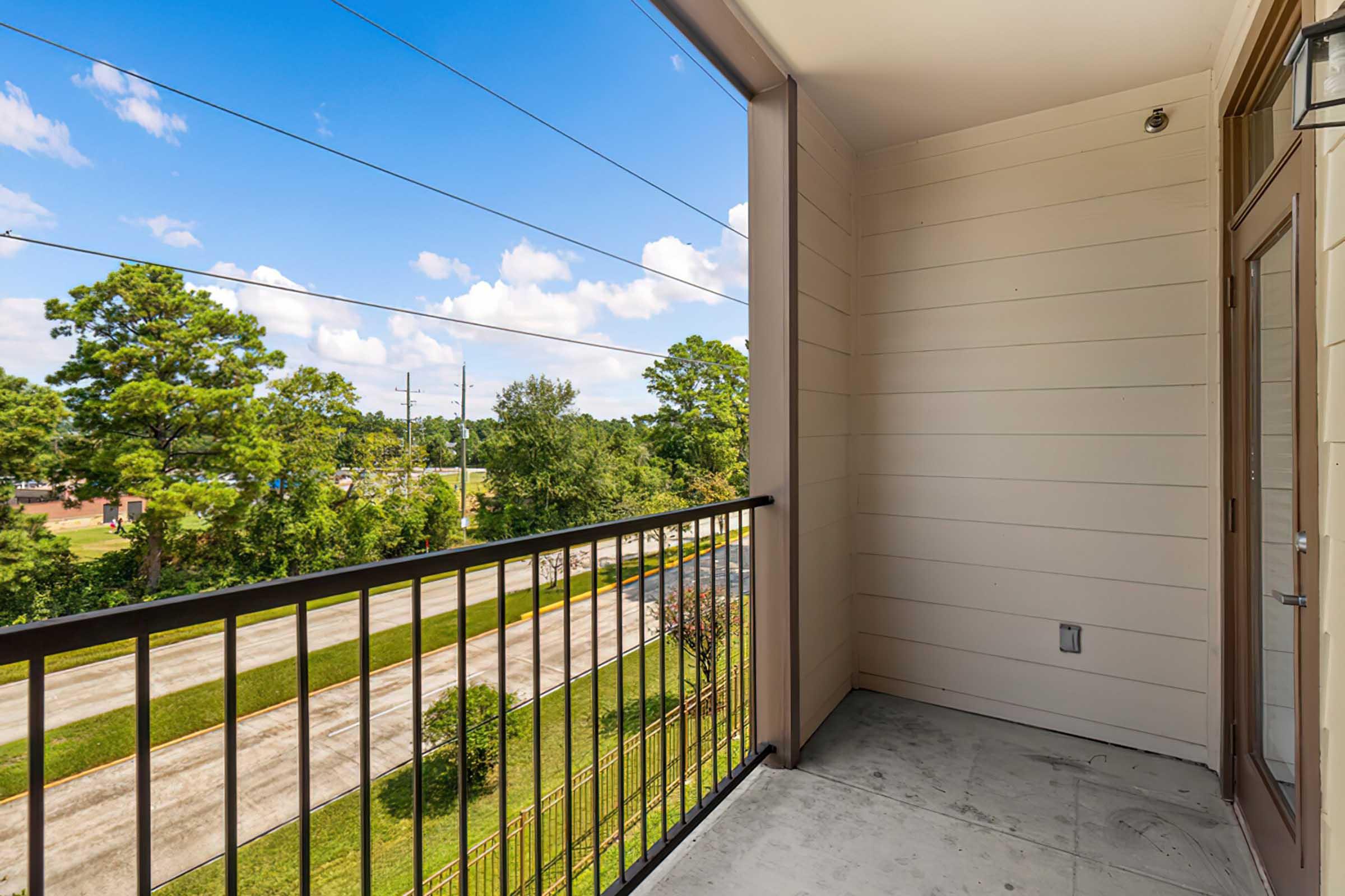 An empty balcony with a metal railing, overlooking a green landscape with trees and a road below. The sky is bright blue with scattered clouds, and the exterior of the building is light-colored with a modern design. Sunlight is casting shadows on the floor of the balcony.