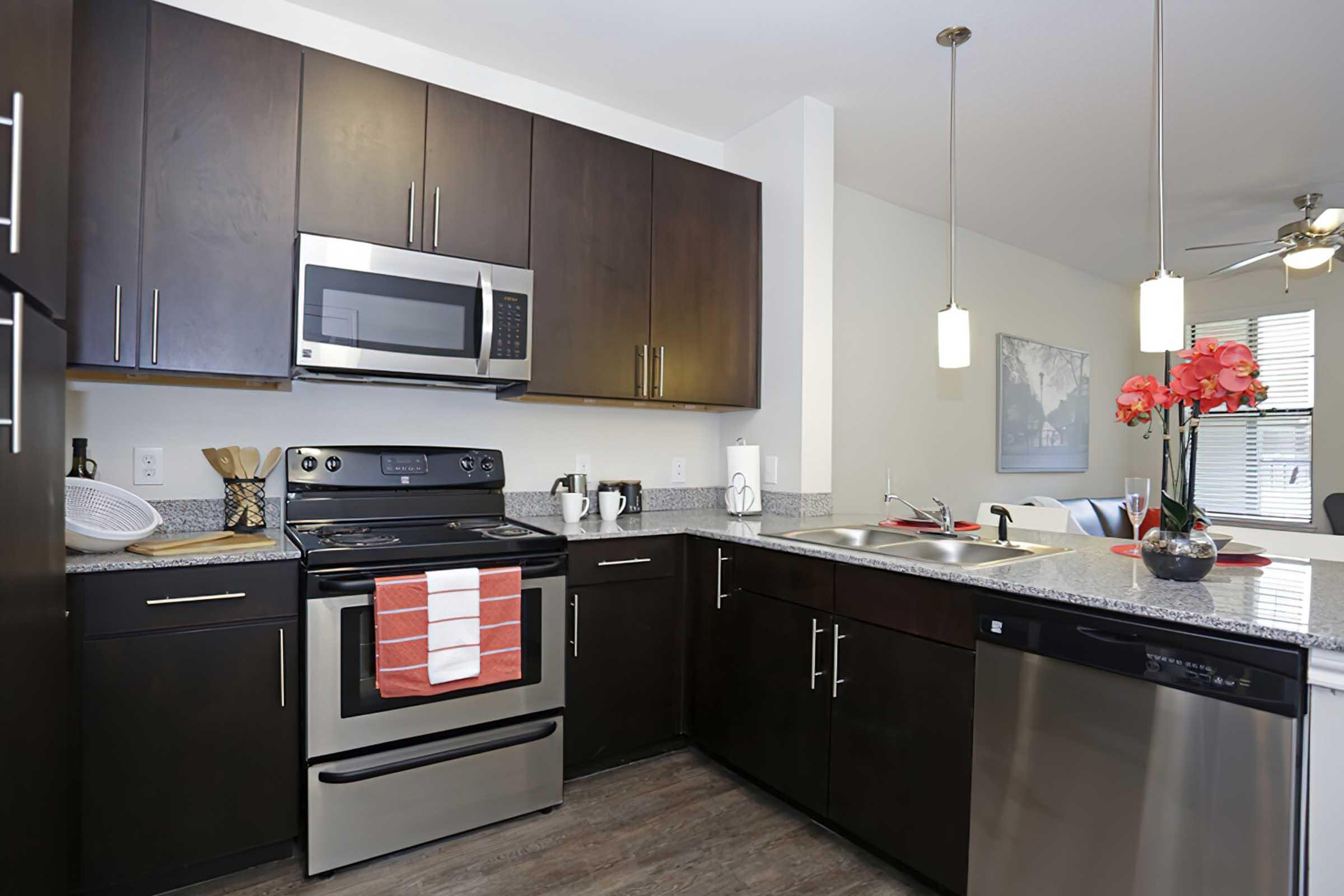Modern kitchen featuring dark wood cabinetry, stainless steel appliances, and granite countertops. The stove has a red and white dish towel hanging from it, and a bowl of utensils is visible. There are pendant lights above the countertop and fresh flowers in a vase, creating an inviting atmosphere.