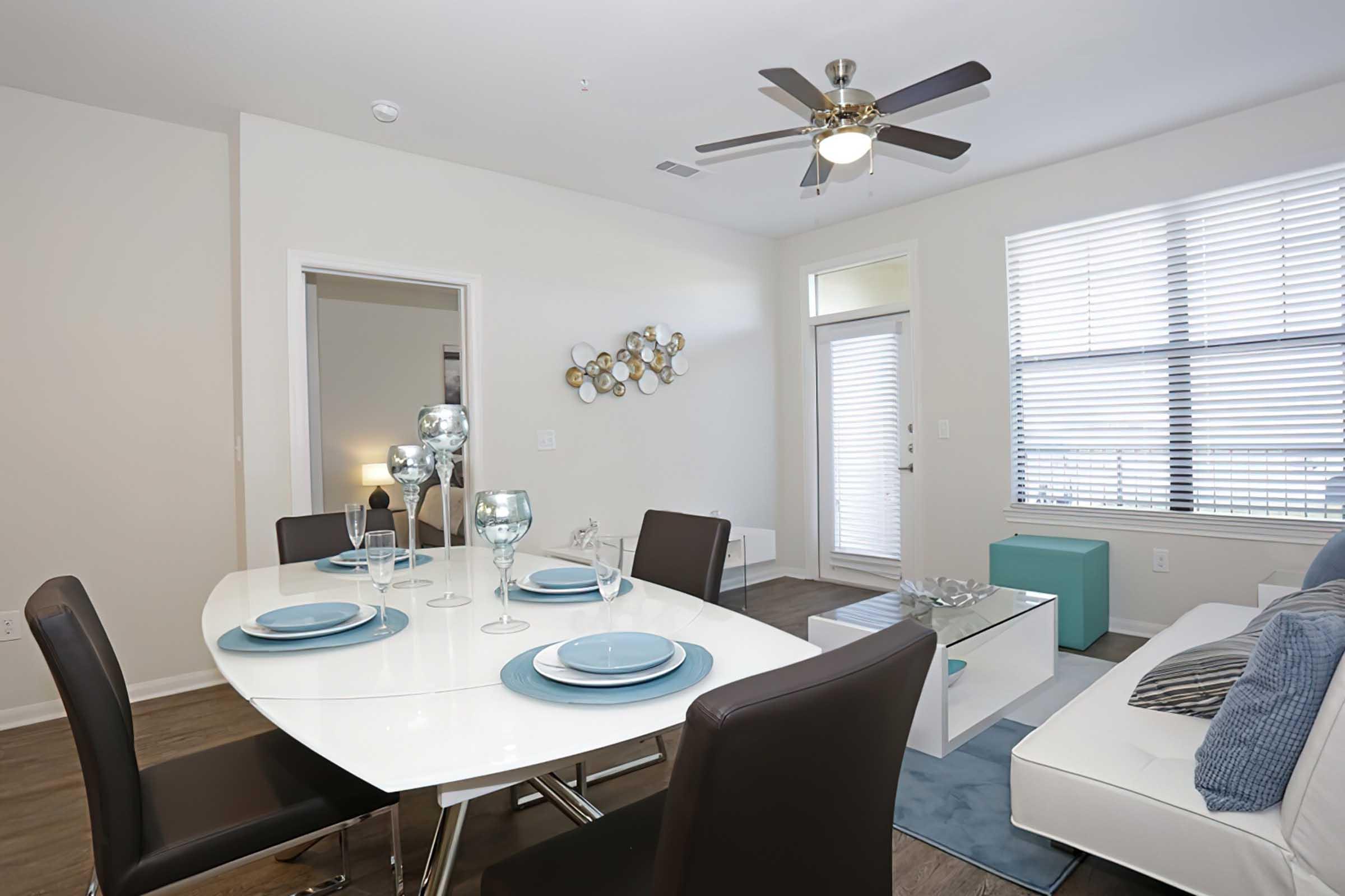 Modern dining area featuring a white oval table set for four with blue plates and glassware. The room includes a light-colored wall, a ceiling fan, and a comfortable living space with a white sofa accented by blue pillows, alongside a stylish decorative piece on the wall. Natural light filters through the window with blinds.