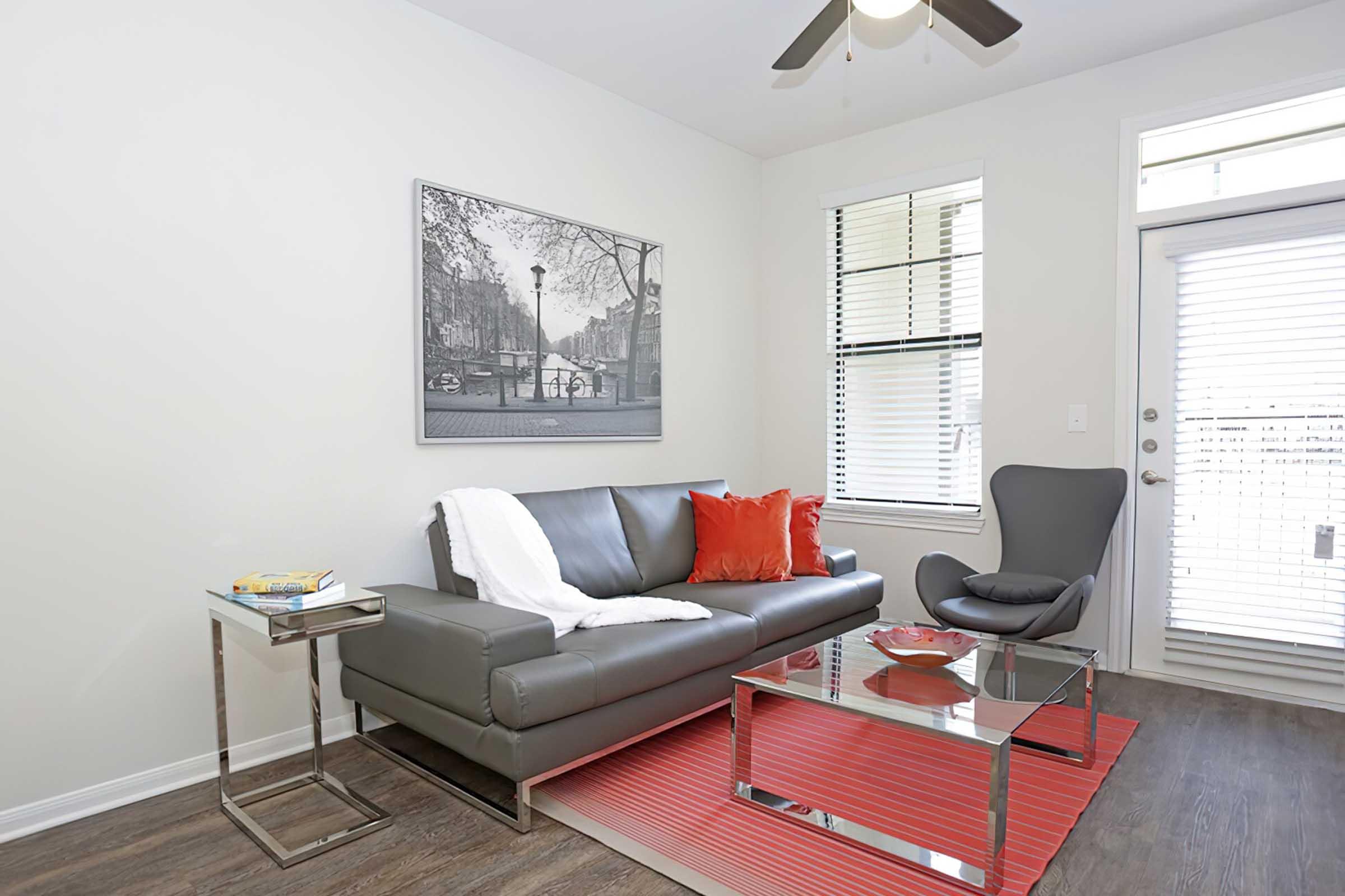 Modern living room featuring a gray leather sofa with red and white accents, a glass coffee table, and a stylish chair. A large black-and-white artwork hangs on the wall, and natural light streams in through a window with blinds. A small side table holds a few books.