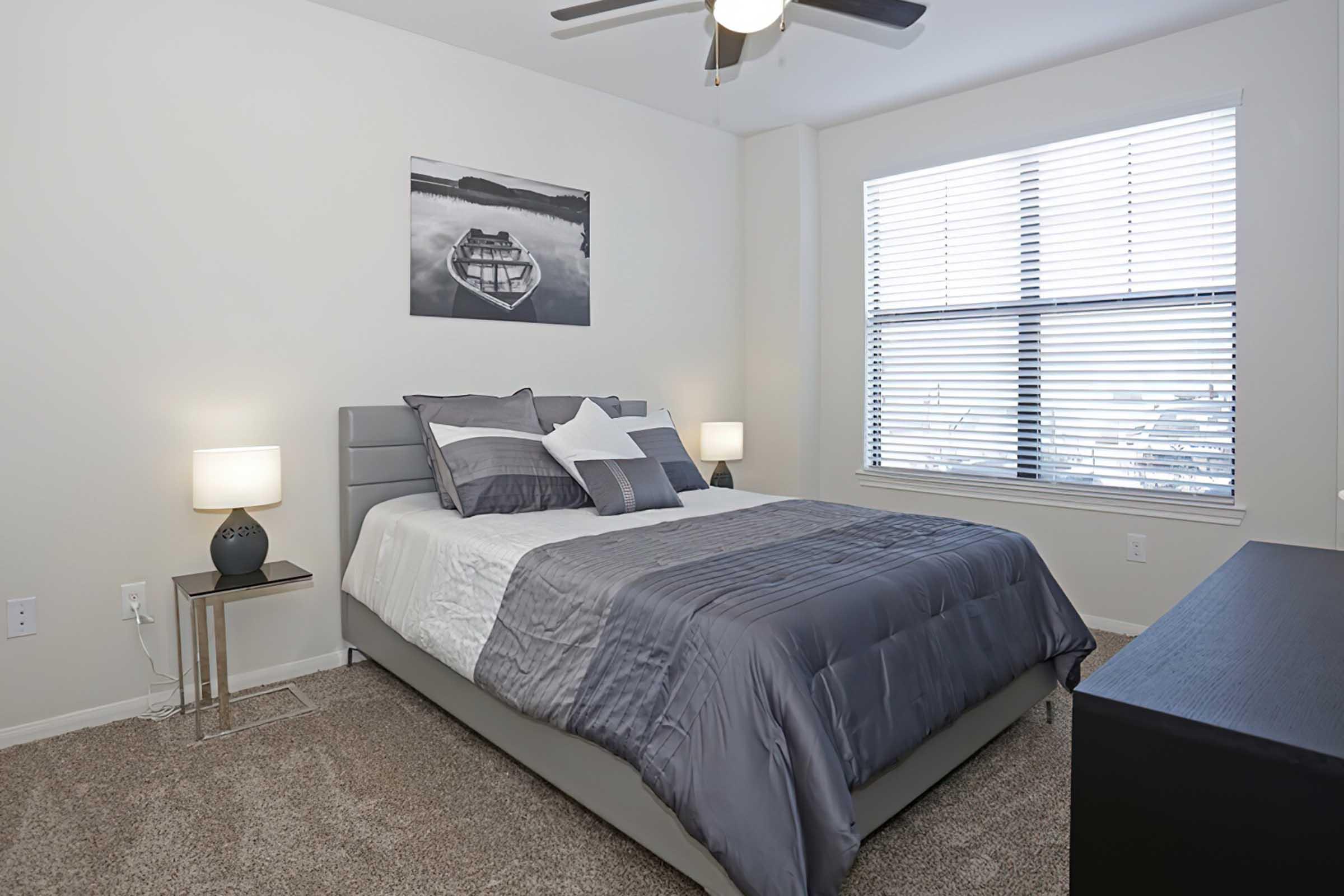 A cozy bedroom featuring a queen-sized bed with gray and white bedding, flanked by modern bedside tables with lamps. A large window allows natural light, adorned with horizontal blinds. A serene black and white landscape photo hangs above the bed, and a dark dresser is positioned against the opposite wall.