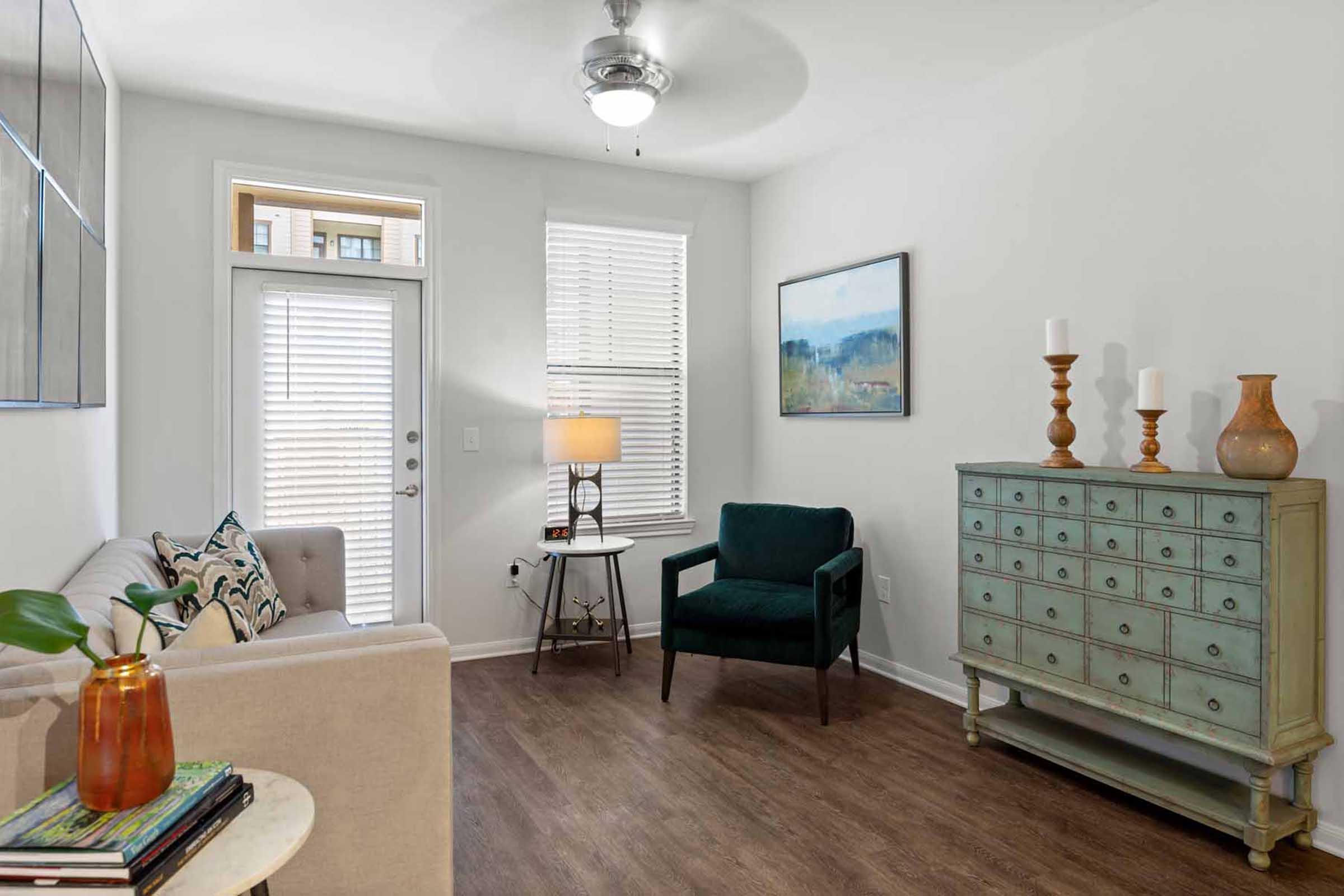A modern living room featuring a light-colored sofa, a green armchair, a wooden side table with a lamp, and a blue dresser. Natural light streams in through window blinds. A piece of artwork hangs on the wall, adding color to the space. The floor is made of wooden planks, creating a warm atmosphere.