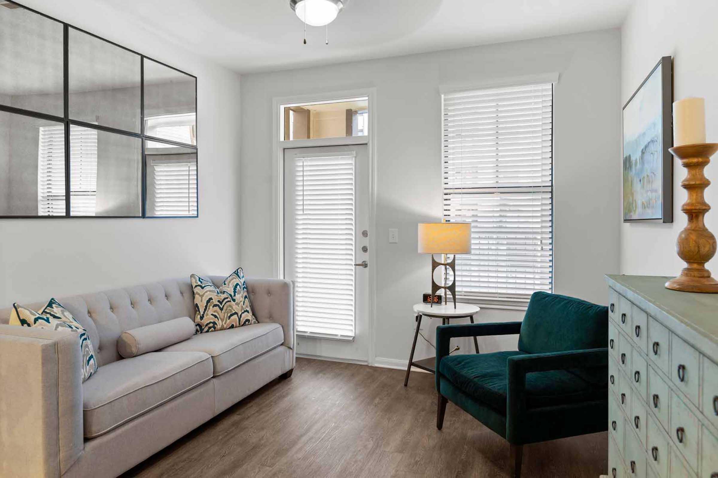 A cozy living room featuring a light gray sofa with decorative pillows, a green armchair, a small round table with a lamp, and a wooden side table. Large windows with blinds allow natural light, and a door leads outside. Minimalistic decor includes a wall art piece and a decorative wooden object.