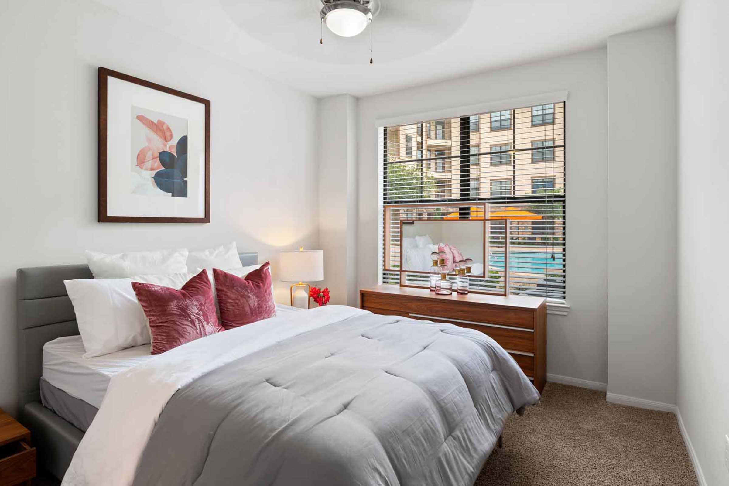 A cozy bedroom featuring a queen-sized bed with gray bedding and plush pink pillows. There is a framed artwork on the wall, a bedside table with a small lamp, and a dresser under a window with wooden blinds, showcasing a view of a pool outside. The room has soft lighting and neutral decor.