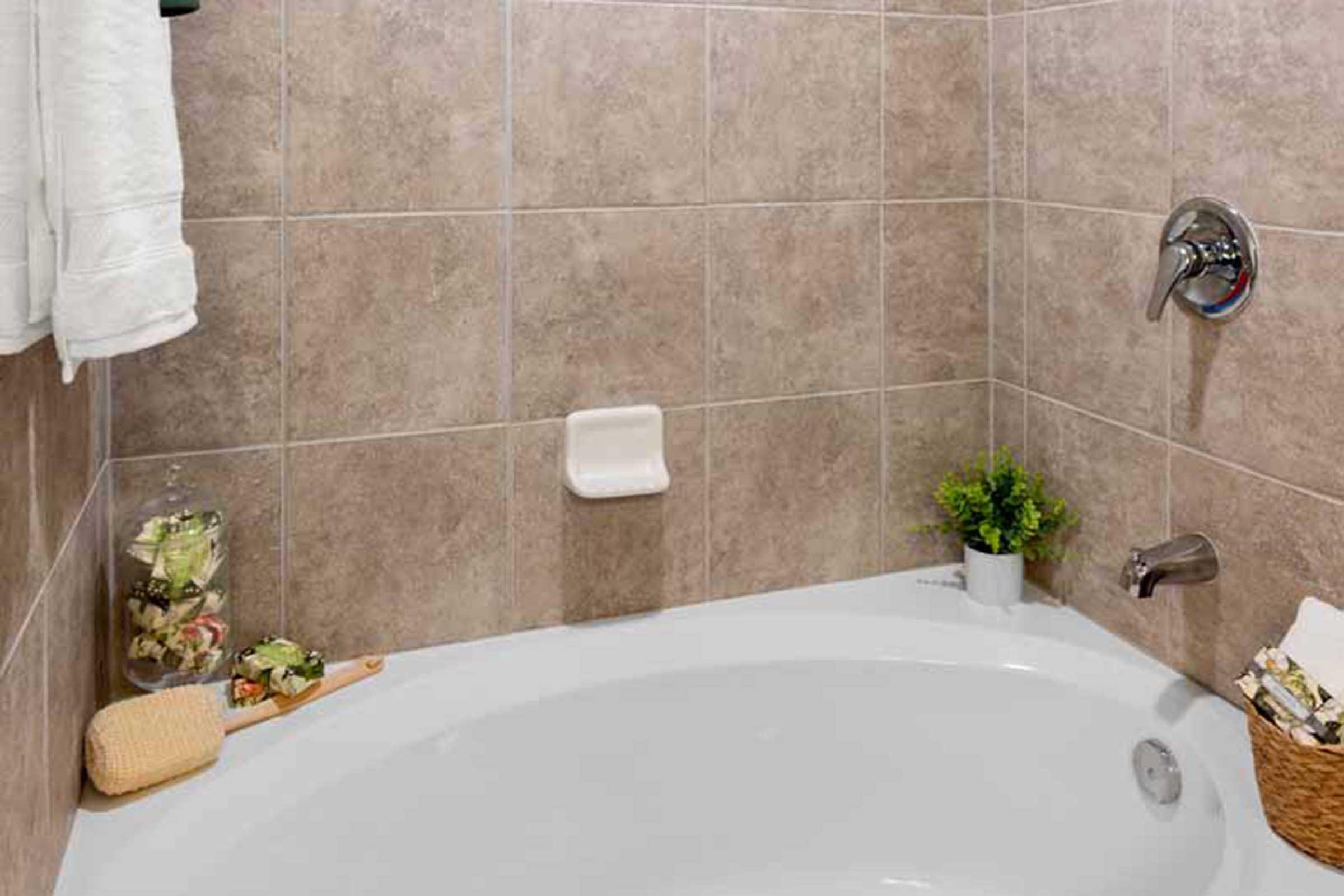 A clean, modern bathroom featuring a white bathtub surrounded by beige tiled walls. There is a towel hanging on a rack, a small potted plant on the edge of the tub, and a jar filled with bath accessories. A shower faucet and a soap dish are also visible, creating a serene and inviting atmosphere.
