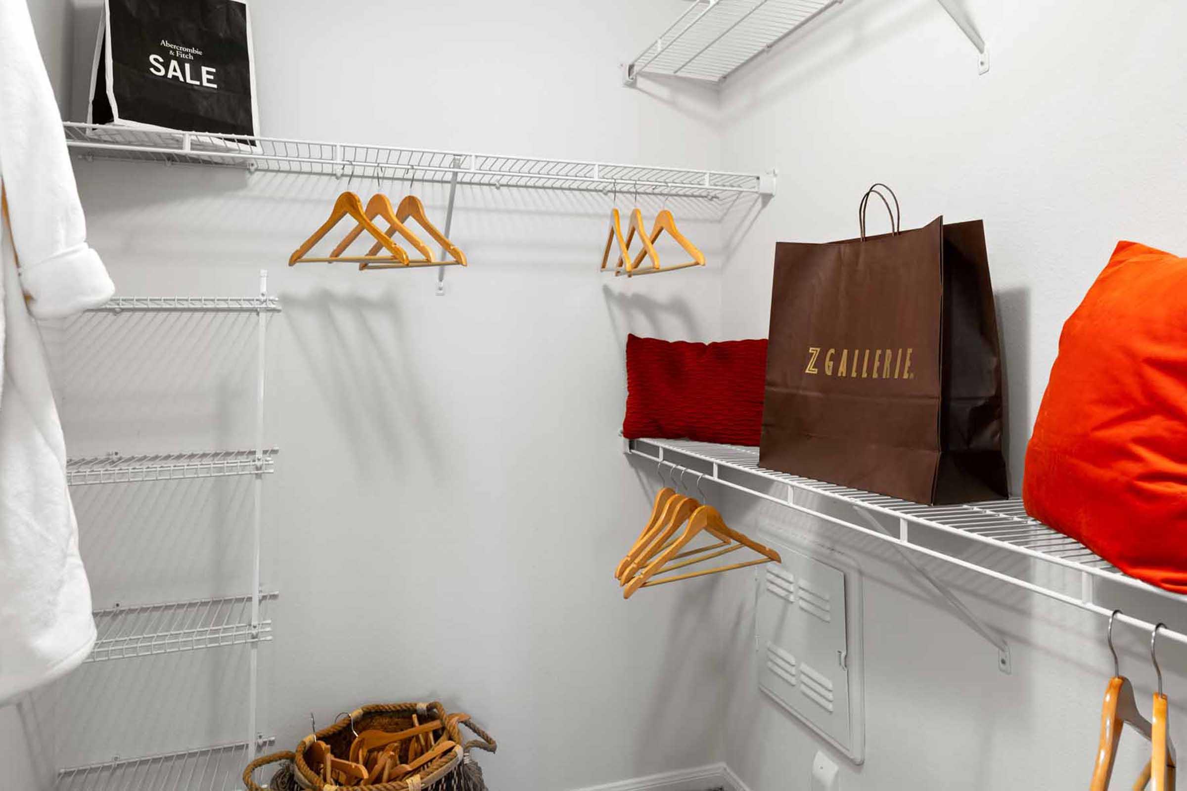 A neatly organized closet featuring metal wire shelving, empty wooden hangers, a large brown shopping bag from Z Gallerie, a red decorative pillow, and a straw basket on the floor. The walls are painted white, enhancing the clean and minimalist look of the space.