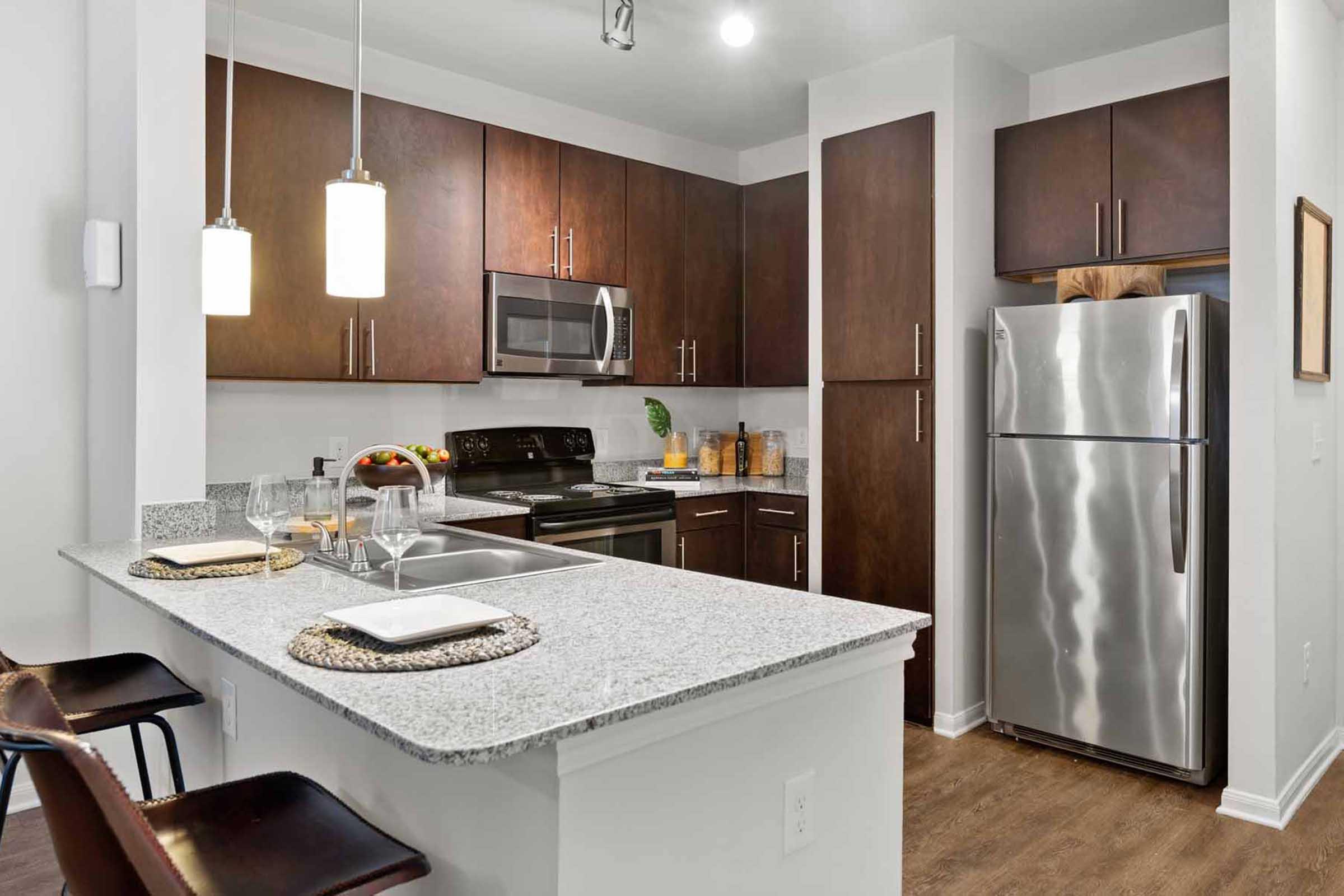 Modern kitchen featuring dark wood cabinetry, stainless steel appliances, and a large granite countertop with bar seating. The space includes an oven, microwave, and a refrigerator, complemented by pendant lighting and decorative items on the counter.