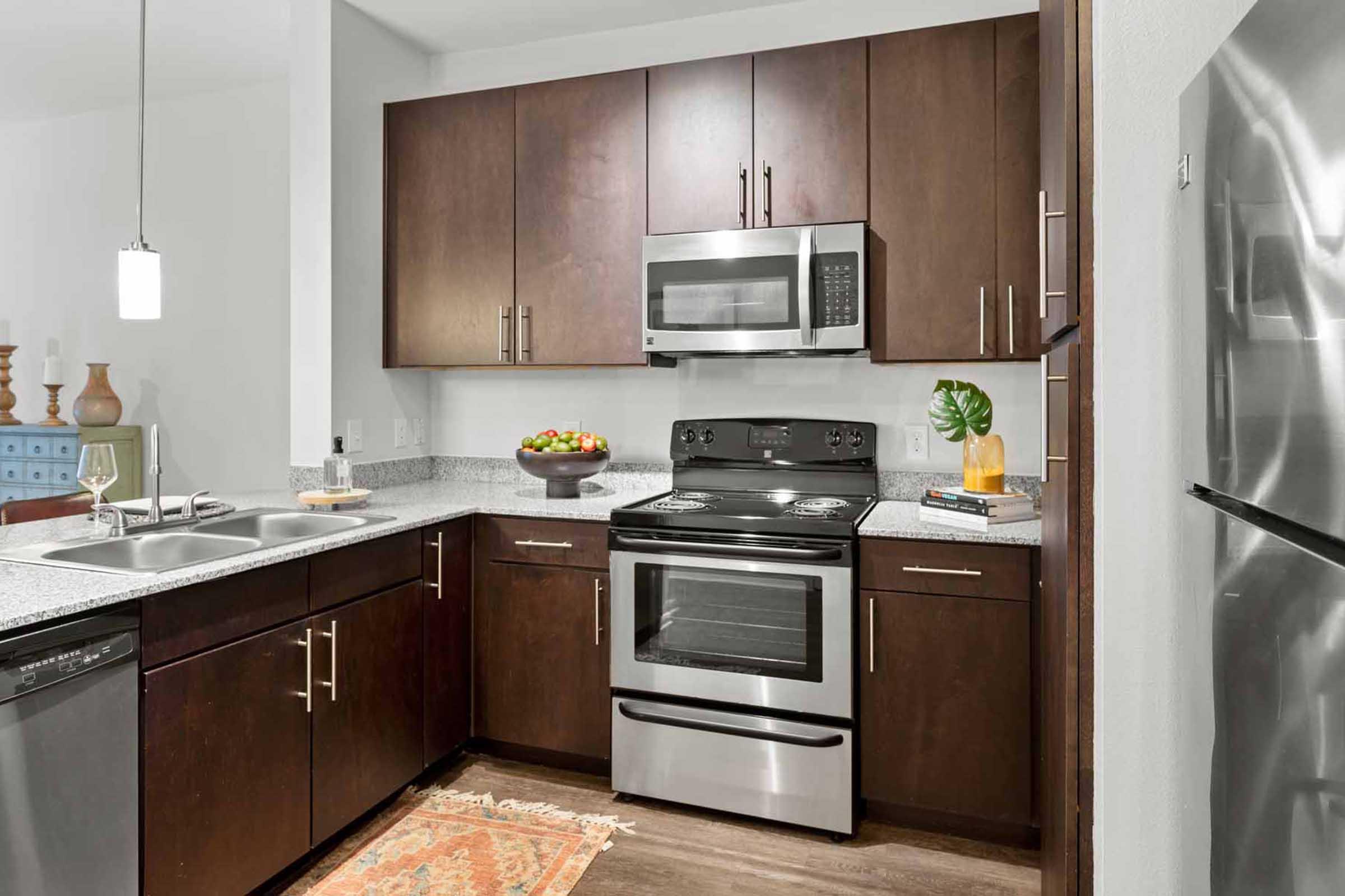 Modern kitchen featuring dark wood cabinets, granite countertops, and stainless steel appliances. Includes a microwave, stove, dual sink, and a colorful fruit bowl. Decorative elements like a small plant and a book on the counter add a cozy touch to the space. A patterned rug is placed on the floor.