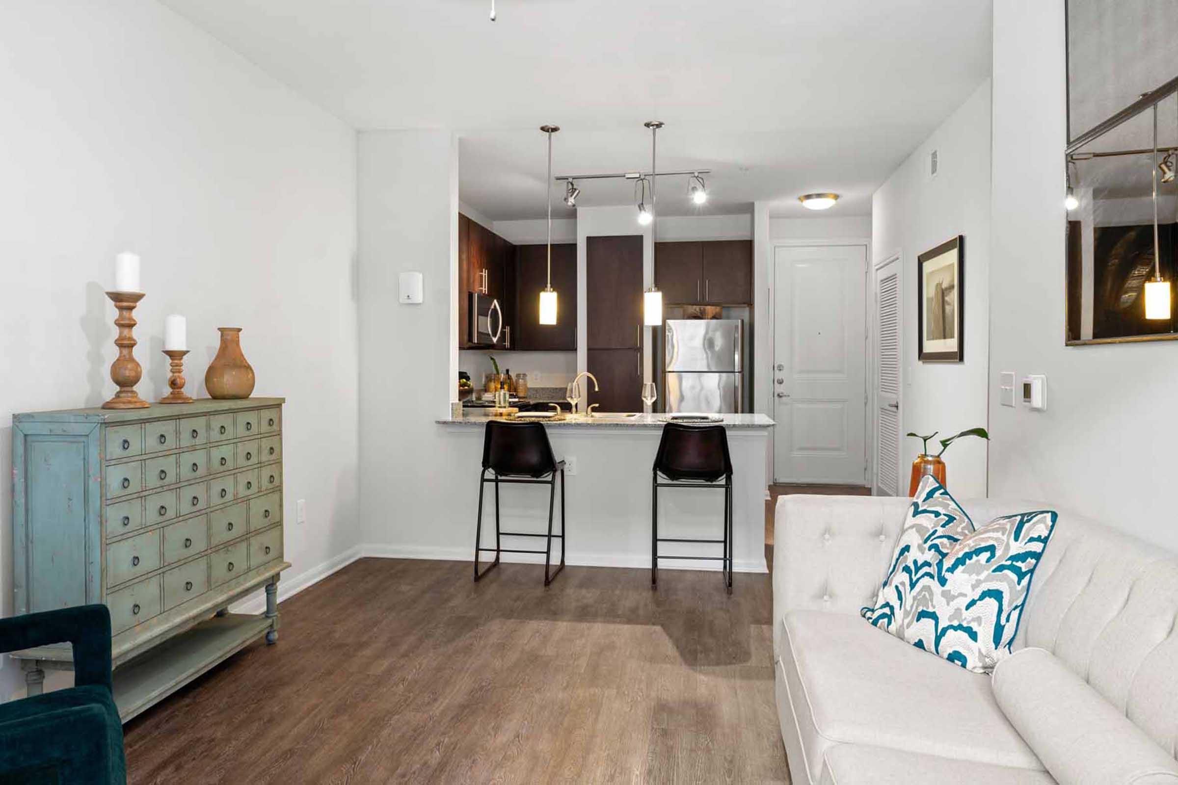 A modern living room with a cream-colored sofa adorned with decorative pillows, featuring a rustic blue dresser and two black bar stools at a kitchen counter. The kitchen has dark cabinetry and stainless steel appliances, while the space is well-lit with natural light and stylish decor elements.