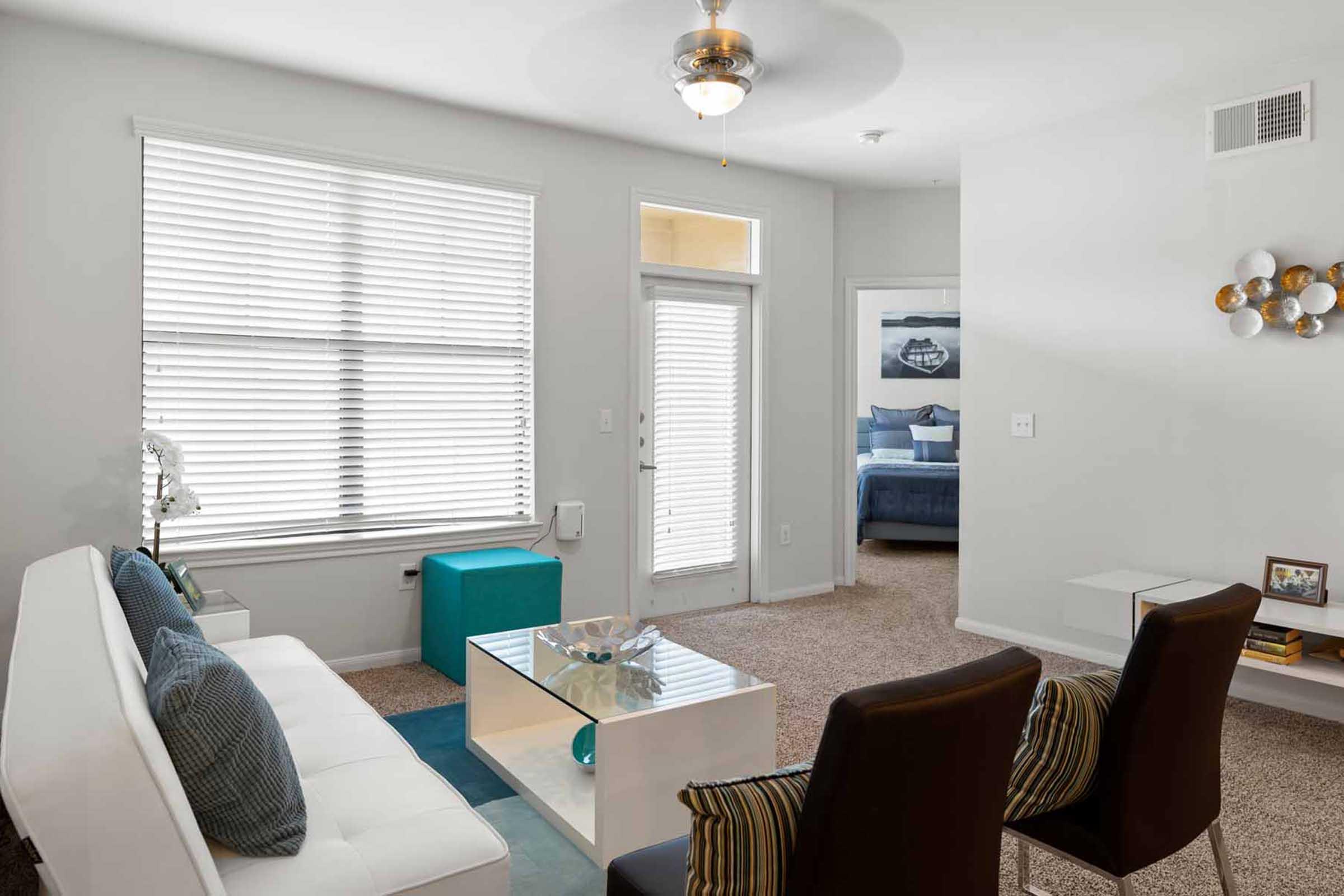 Bright and modern living room featuring a white sofa with blue accent pillows, a glass coffee table, and two stylish chairs with striped cushions. Natural light floods the space through large windows with blinds. A doorway leads to a bedroom with a blue bedspread visible in the background.