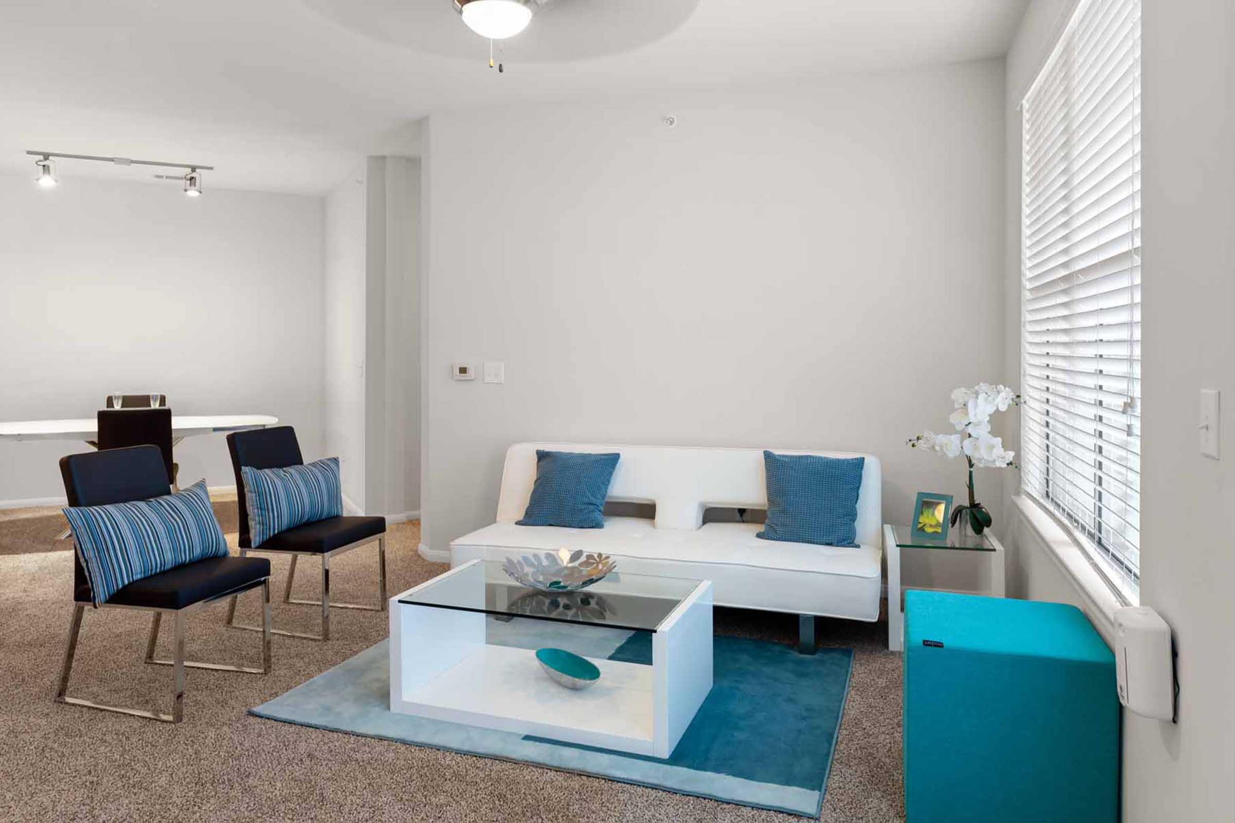 A modern living room featuring a white sofa with blue cushions, two black chairs with blue striped backs, a glass coffee table, and a decorative bowl. The space has neutral walls, a light carpet, and a small turquoise cabinet. A large window provides natural light, and there are potted plants adding greenery.