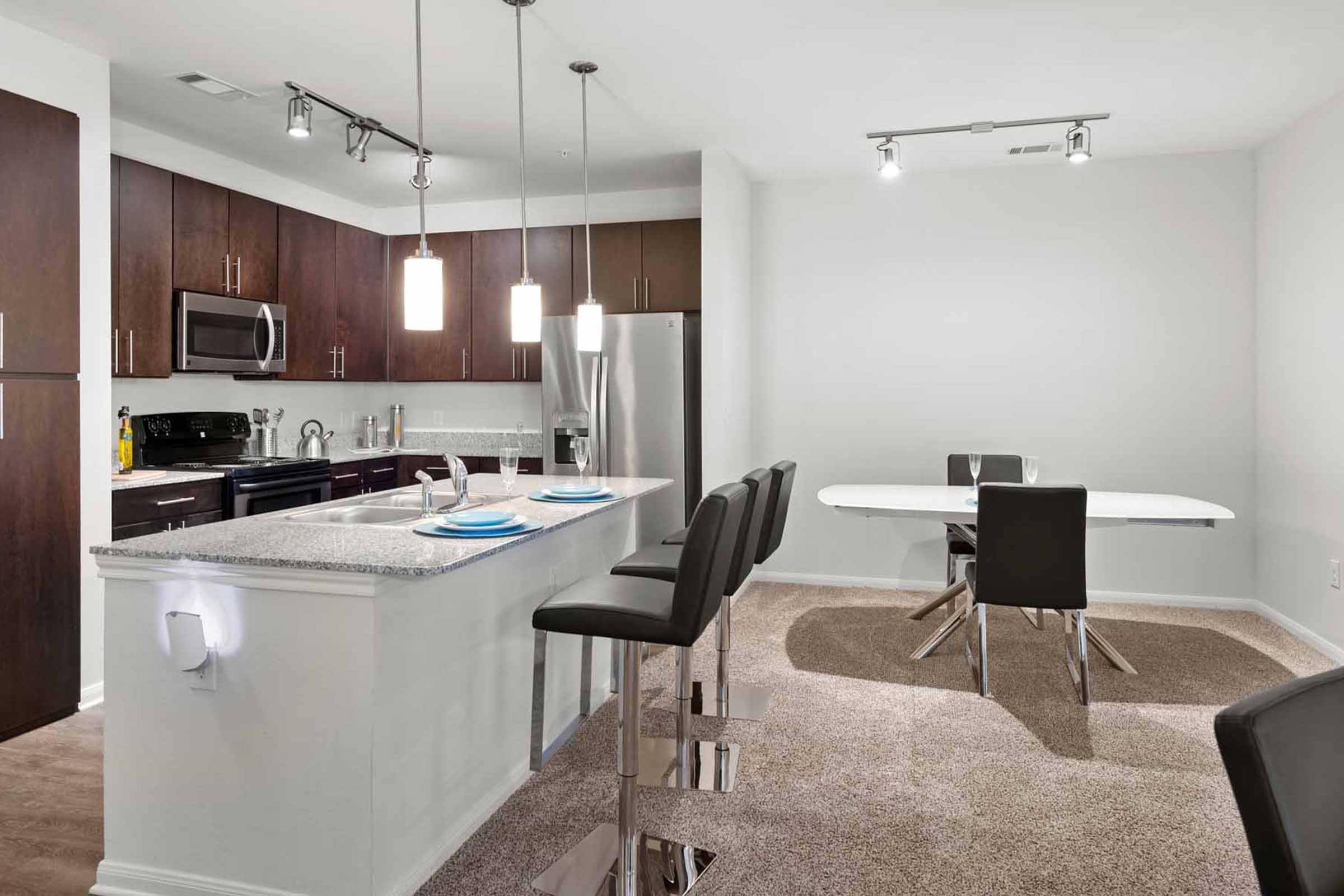Modern kitchen with dark wood cabinets and stainless steel appliances. A granite island with two bar stools faces the dining area, which features a white table with four chairs. Soft lighting from pendant lights enhances the contemporary design. The floor is carpeted, adding warmth to the space.
