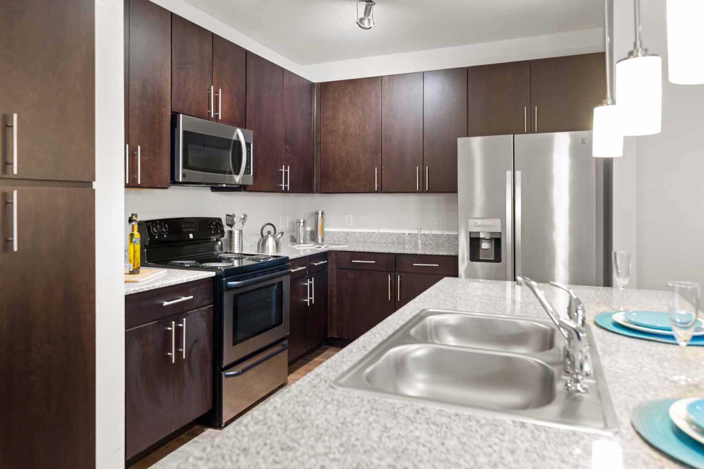 Modern kitchen featuring dark wooden cabinets, stainless steel appliances including a microwave, oven, and refrigerator, a double sink with a sleek faucet, and a granite countertop. There are decorative items on the counter and plates set on the surface, enhancing the contemporary aesthetic.