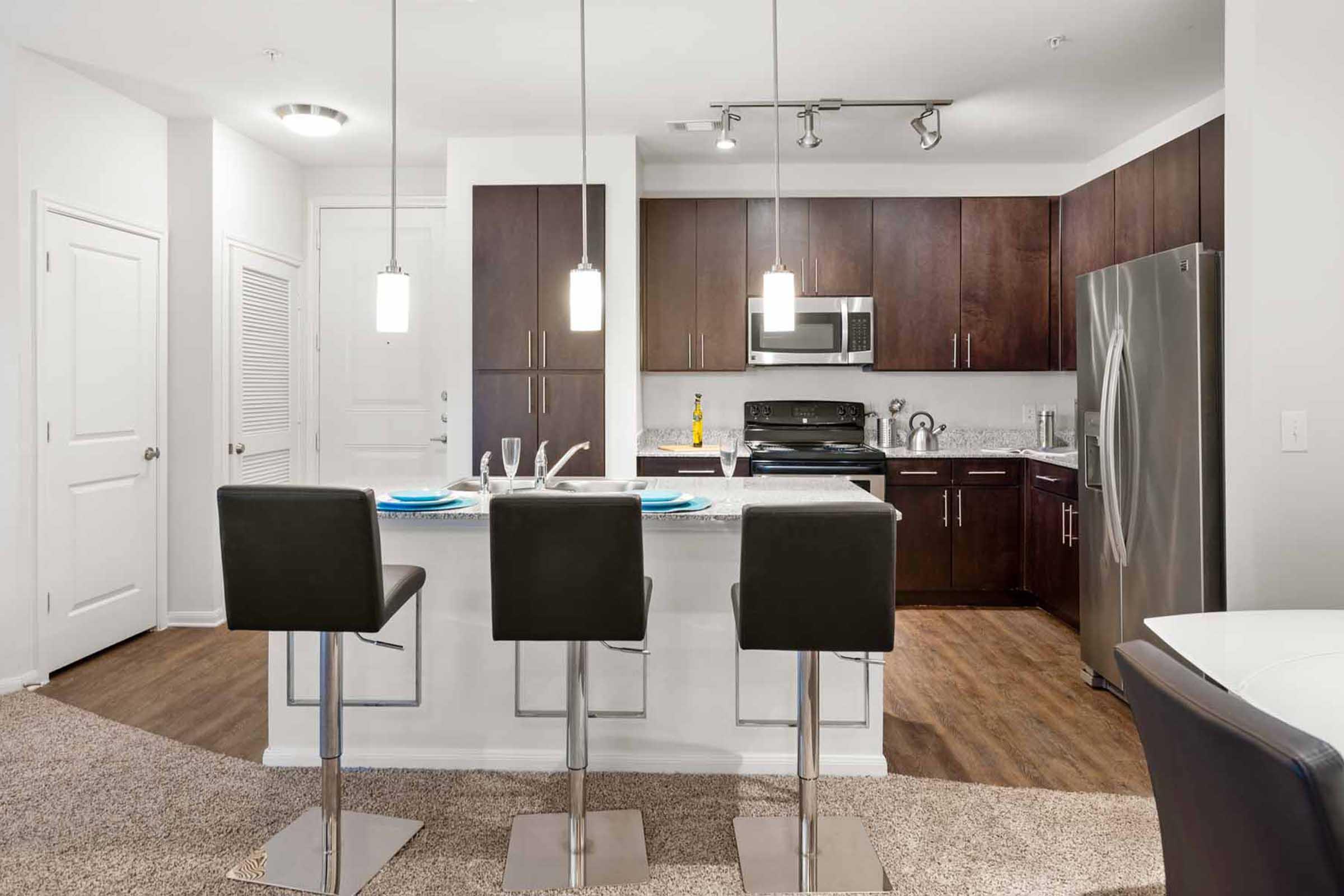 Modern kitchen featuring dark wood cabinetry, stainless steel appliances, and a spacious island with three black bar stools. The area has sleek pendant lighting, a light-colored countertop, and a cozy carpeted living space nearby. Doors lead to additional rooms, creating an open-concept design.