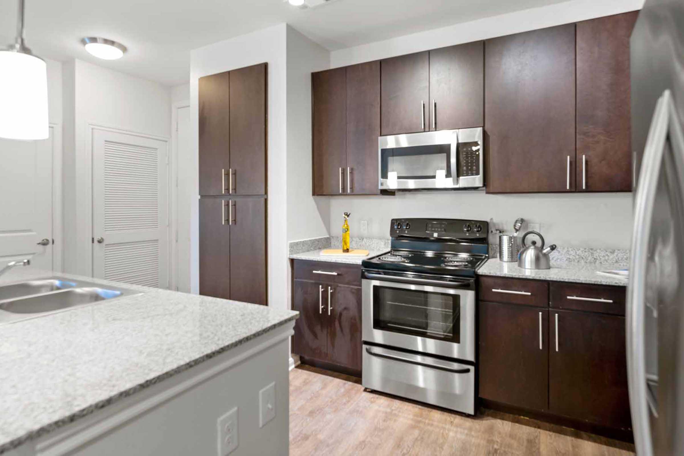 Modern kitchen featuring dark wood cabinetry, a stainless steel stove, microwave, and refrigerator. A countertop with a sink is visible in the foreground, and the space is well-lit with overhead lighting. Gray granite countertops complete the contemporary design.