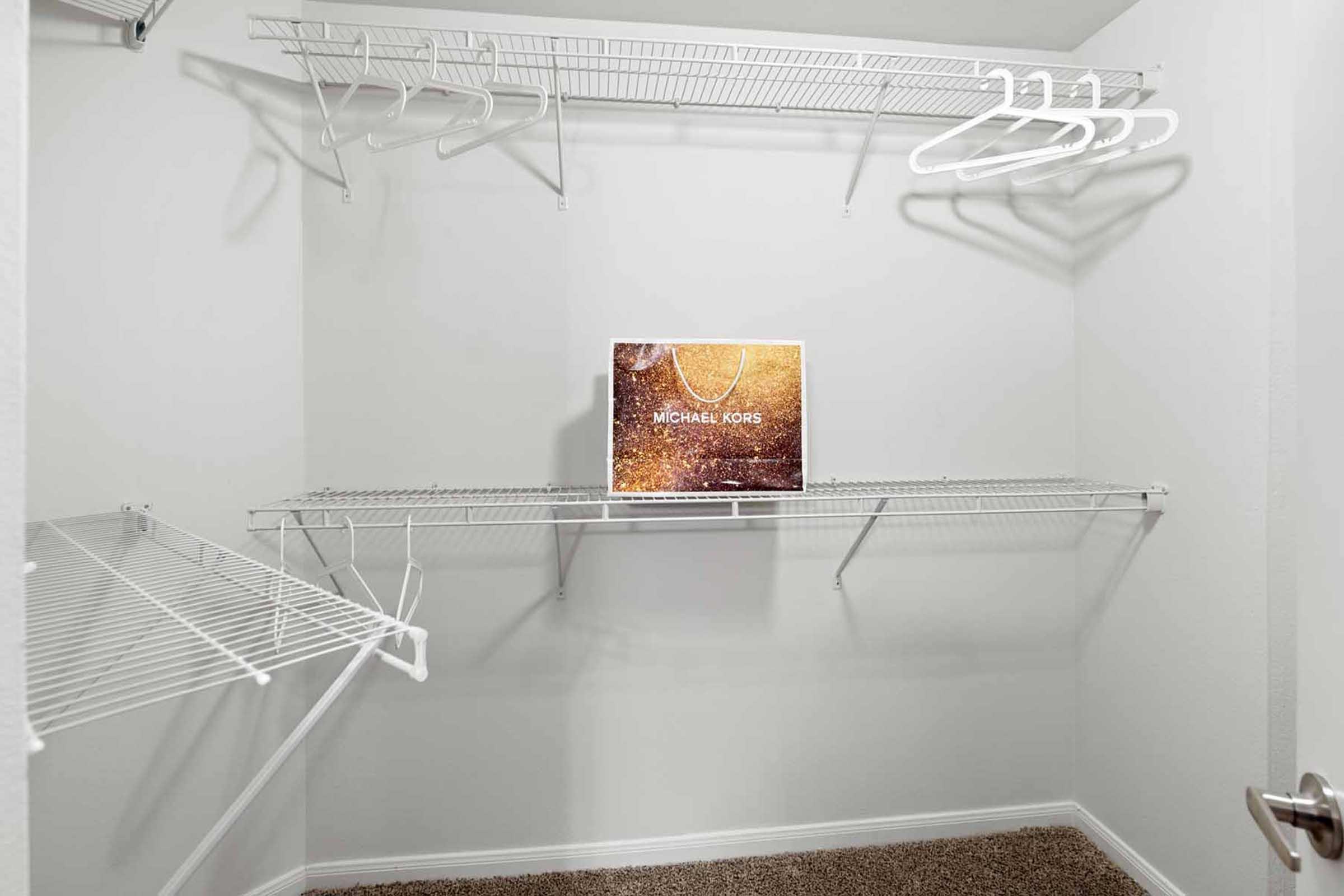An empty closet featuring white wire shelves and hangers. A Michael Kors box is placed on one of the shelves, adding a touch of style to the otherwise minimalistic space. The walls are plain white, and the floor is carpeted.