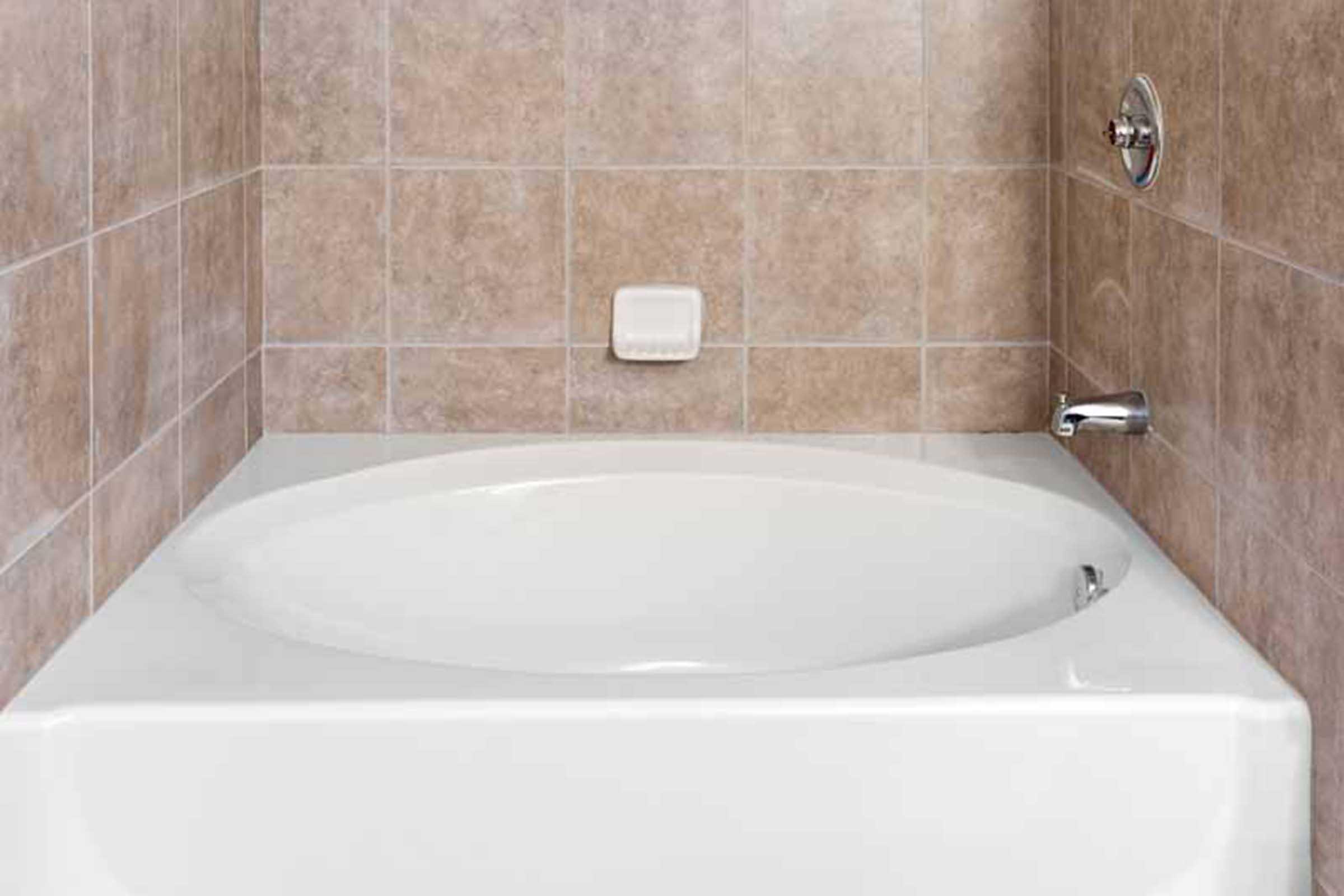 A modern bathroom featuring a white, oval bathtub with a chrome faucet, set against a tiled wall in light brown tones. The bathtub is empty, showcasing its clean design, with a soap dish mounted on the wall nearby.