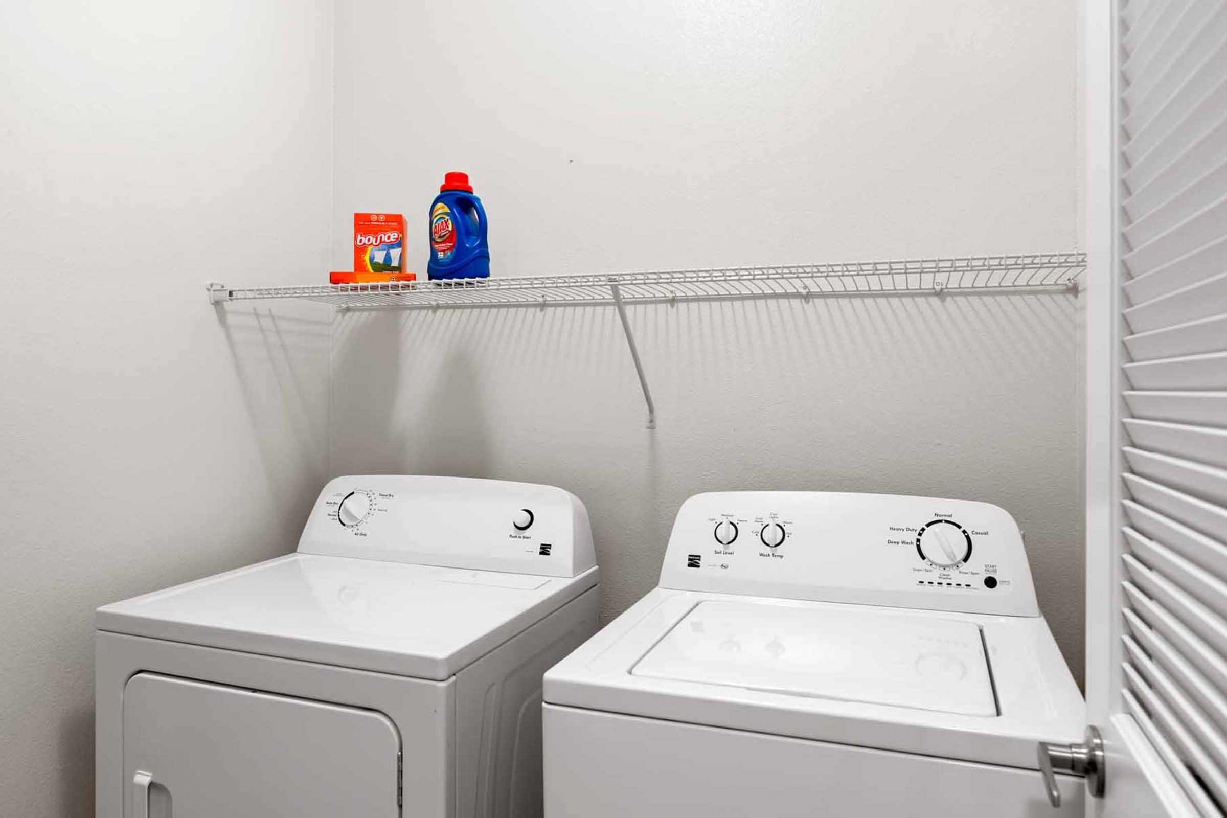 A laundry room featuring a white washing machine and a dryer side by side. Above them, a wire shelf holds a box of laundry detergent pods and a bottle of liquid laundry detergent. The walls are painted in a neutral color, creating a clean and organized space.