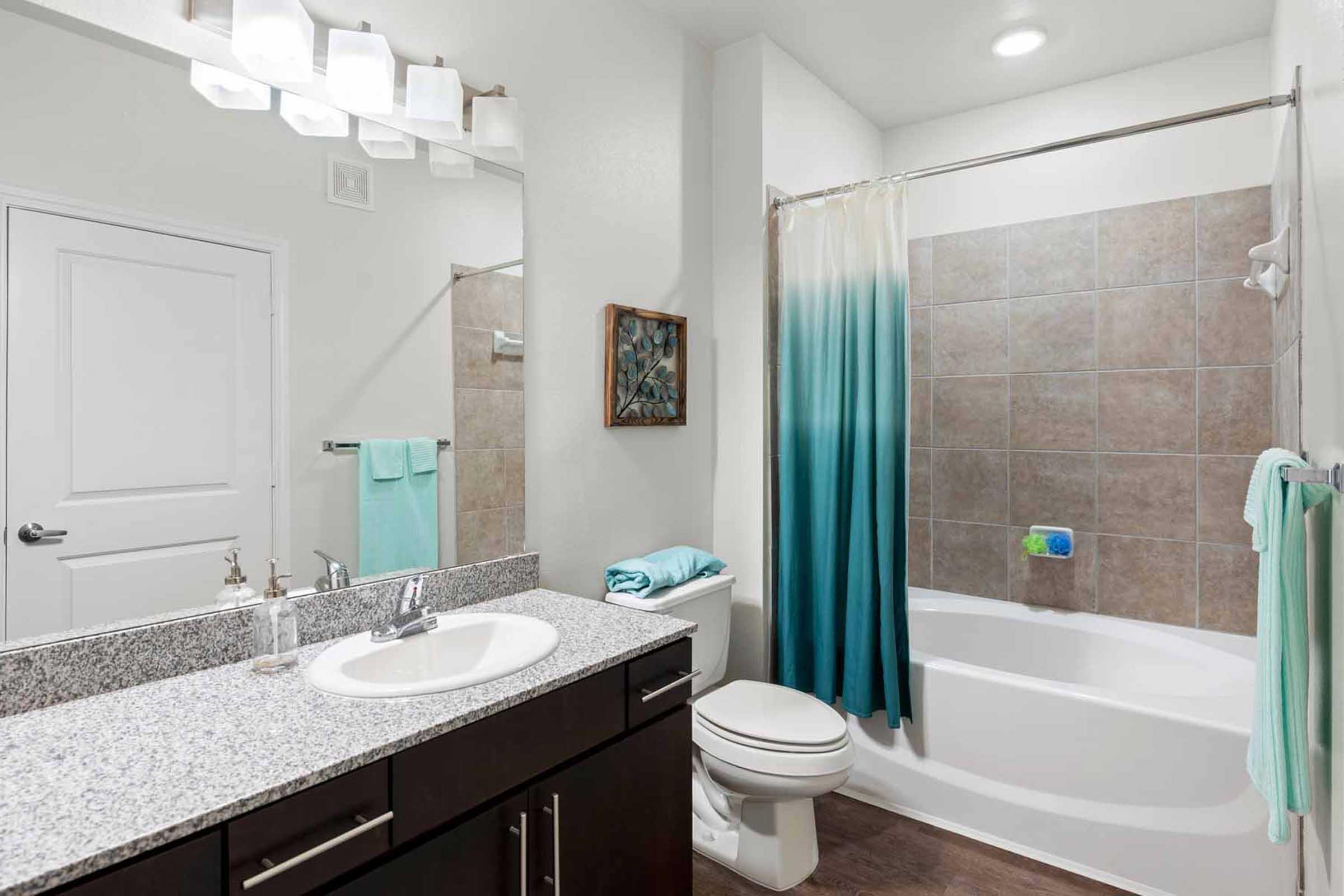 A modern bathroom featuring a bathtub with a shower curtain that fades from light blue to teal. The space includes a granite countertop with a sink, a large mirror, and light fixtures above. Soft turquoise towels hang nearby, and there is a small piece of artwork on the wall. The overall design is clean and contemporary.