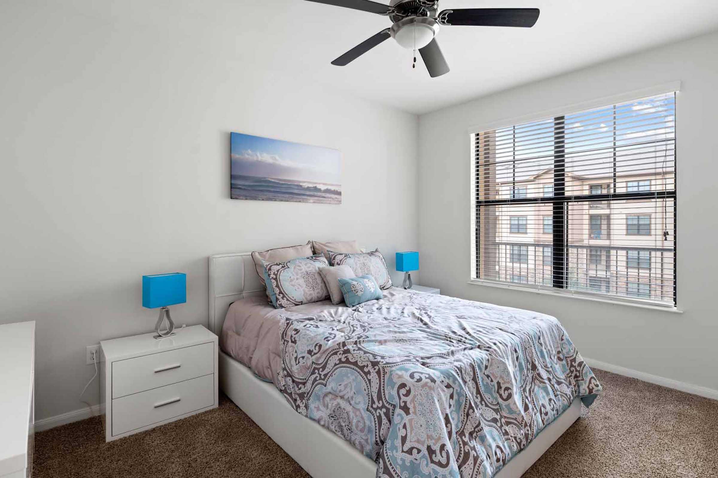 A modern bedroom featuring a cozy bed with patterned bedding, two bedside tables with blue lamps, and a window with blinds allowing natural light. A piece of wall art depicting a scenic view adds a decorative touch to the light-colored walls. The room has a soft carpeted floor.