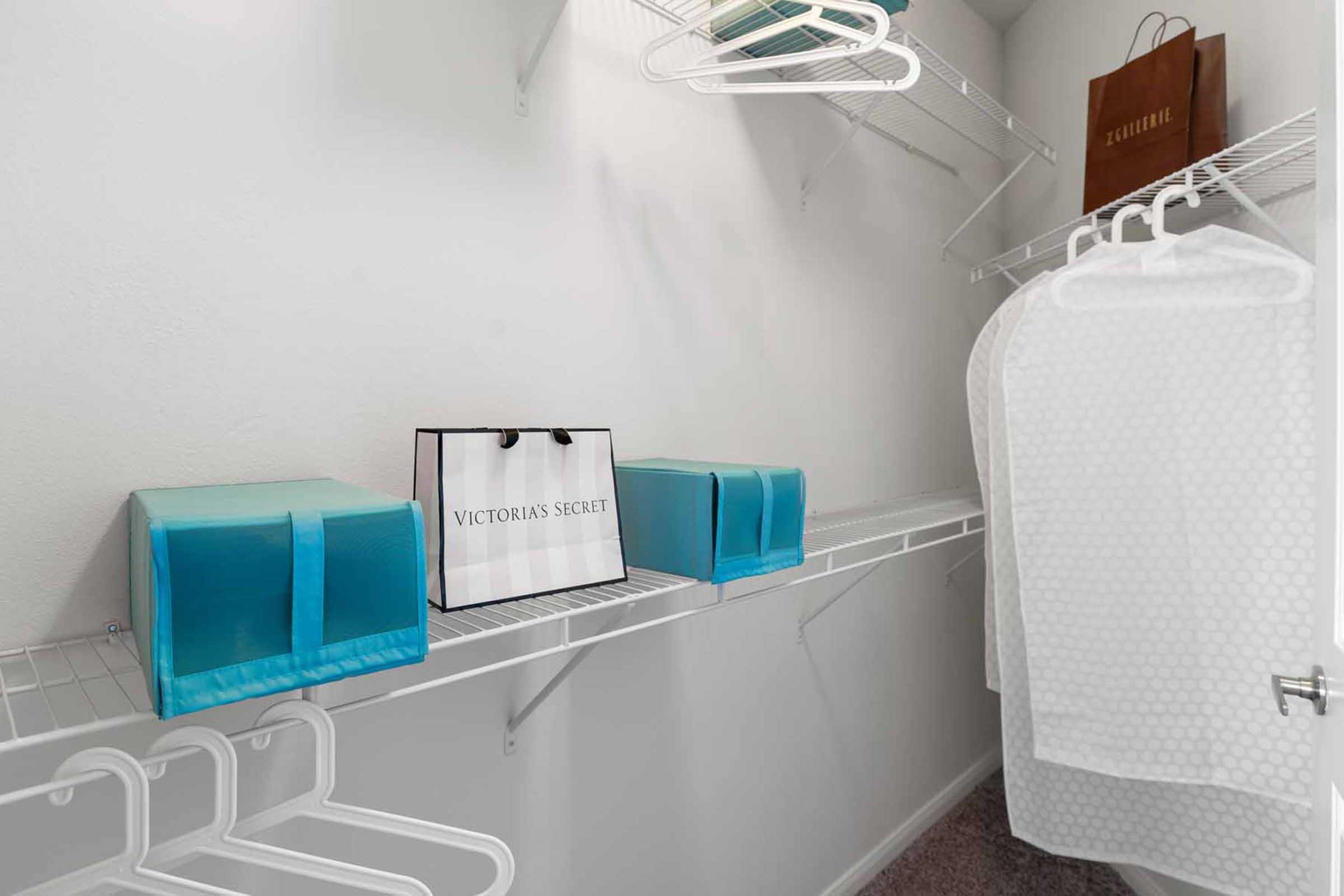 A brightly lit closet featuring white wire shelves. On the shelves, there are two blue storage boxes, a black shopping bag labeled "Victoria's Secret," and several white hangers. The walls are plain and the flooring is carpeted.