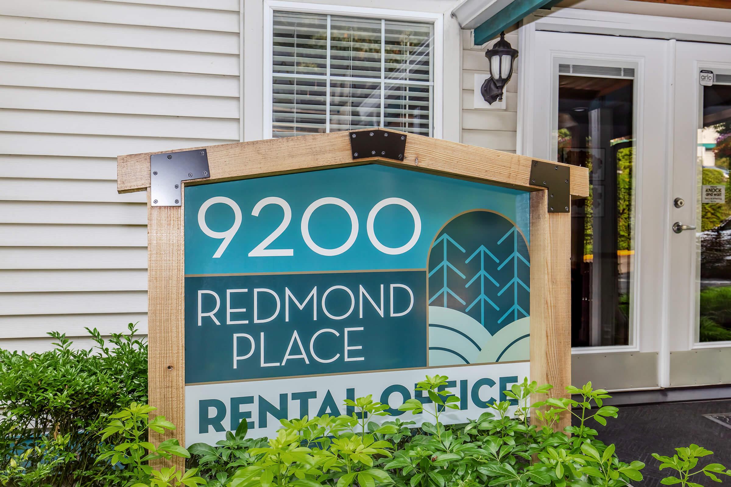 Sign for Redmond Place featuring the address "9200" and labeled "Rental Office." The sign is wooden with a modern design, situated in front of a building with a green landscape around it. There are large windows visible in the background indicating an inviting entrance.