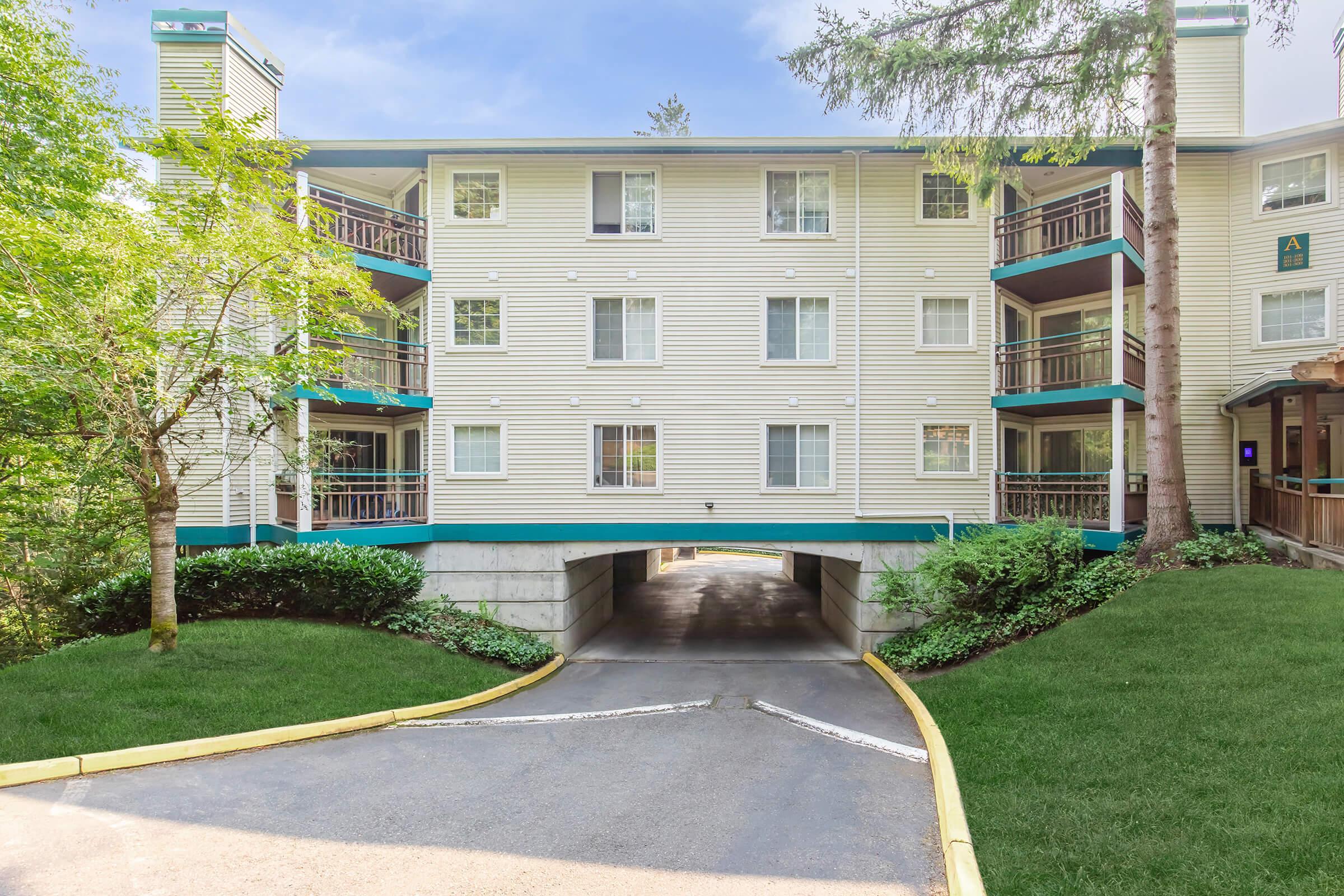 A three-story residential building with a light-colored exterior and green accents. The structure has multiple balconies and a covered parking area underneath. Surrounding the building are trees and well-maintained grass, creating a serene and inviting atmosphere. The sky is clear and bright.