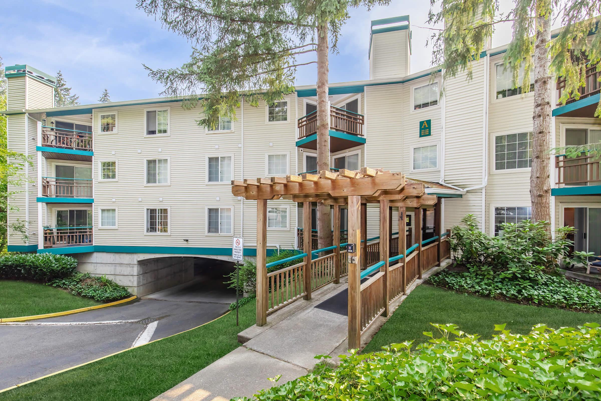 A multi-story residential building with white siding and green trim. The entrance features a wooden pergola and is surrounded by well-maintained landscaping, including shrubs and grass. The driveway leads under the building, providing access to parking. Tall trees are visible nearby, adding to the greenery.