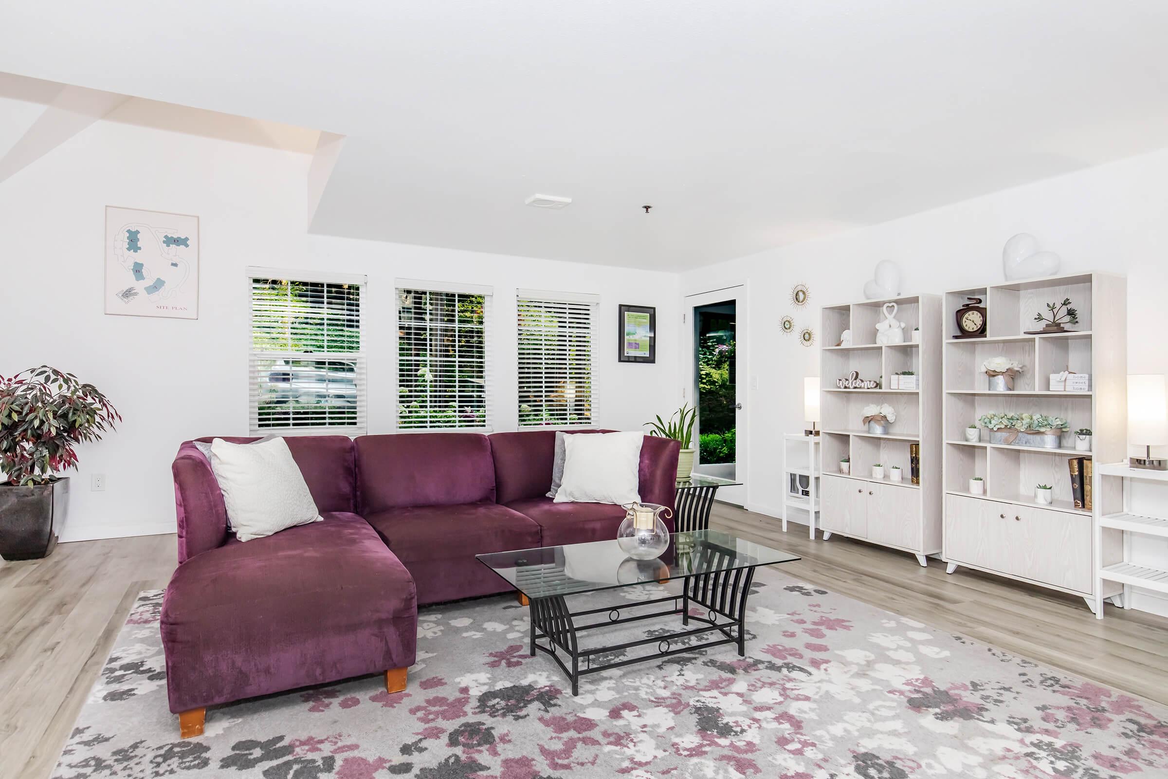 A cozy living room featuring a purple sectional sofa, a glass coffee table, and a light-colored bookshelf displaying decorative items. Large windows allow natural light, and a floral area rug adds a touch of color. Indoor plants enhance the inviting atmosphere.