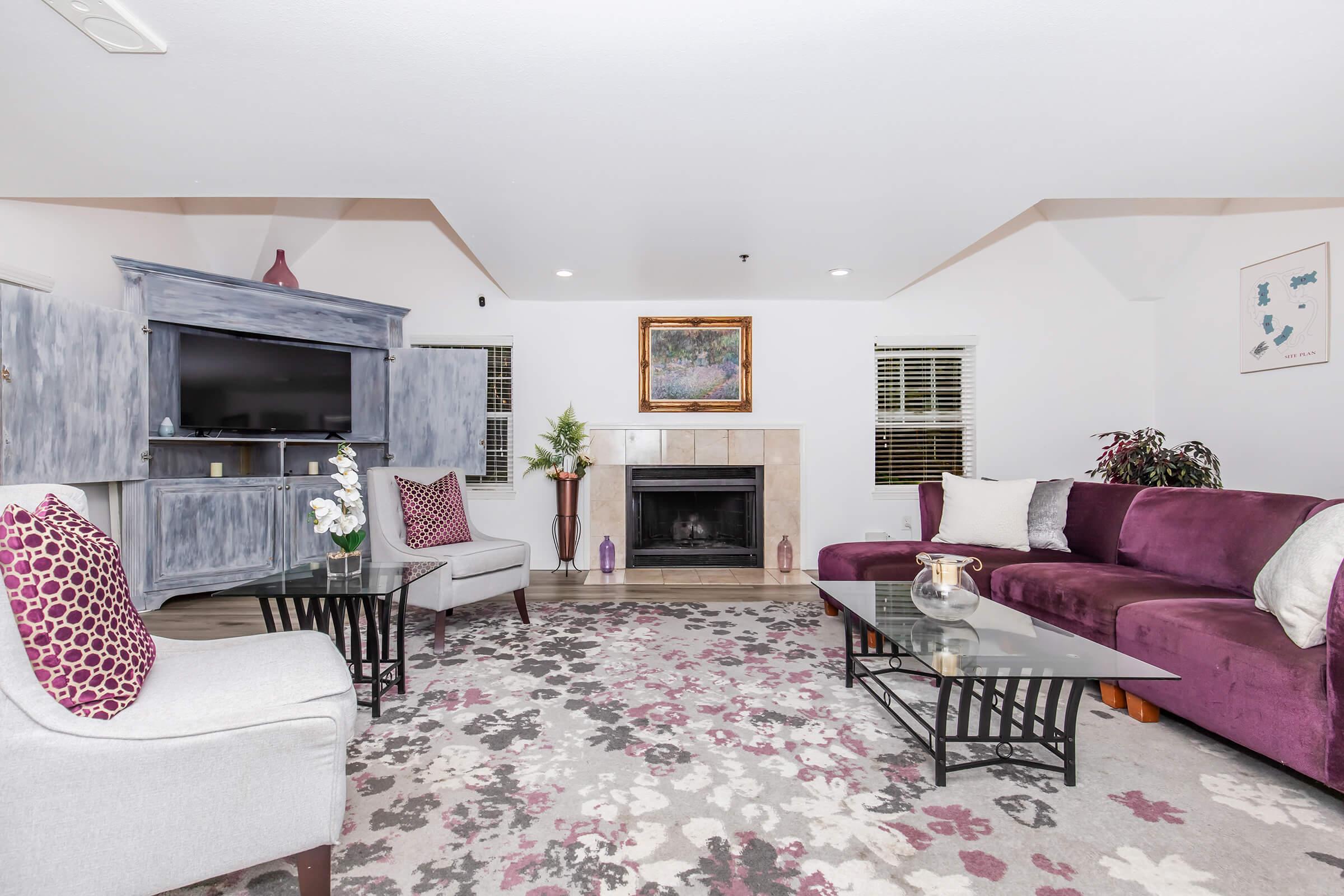A cozy living room featuring a purple sectional sofa and two light-colored chairs. A glass coffee table sits on a floral-patterned rug, while a fireplace is centered against the wall. There’s a blue cabinet with a television, decorative plants, and a framed picture above the fireplace, creating a warm and inviting atmosphere.