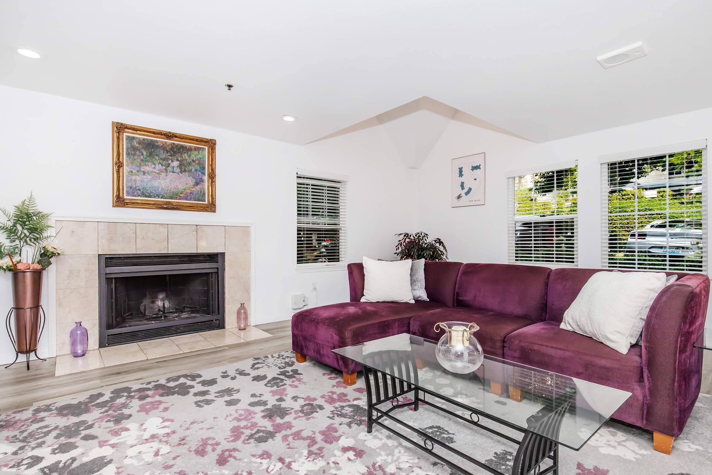 A cozy living room featuring a purple sectional sofa with decorative pillows, a glass coffee table, and a fireplace. There are large windows with natural light and a colorful area rug on the floor. A framed artwork hangs on the wall, and indoor plants add a touch of greenery.