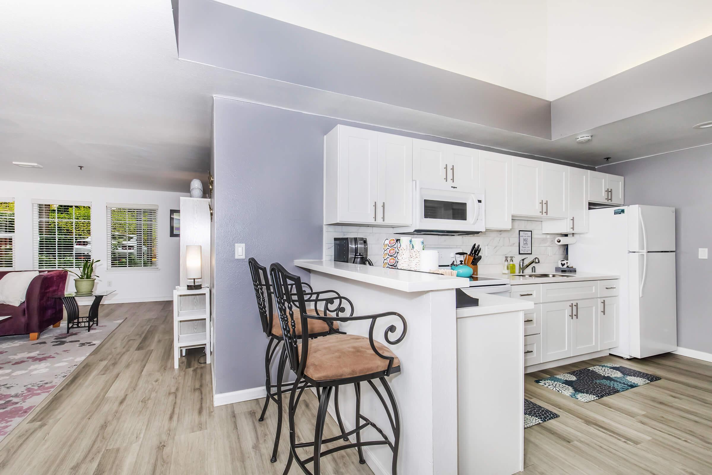 Modern kitchen with white cabinets and a breakfast bar featuring three stools. There is a microwave, refrigerator, and various kitchen utensils on the counter. In the background, a living area with a purple chair and large windows is visible, allowing natural light to brighten the space.